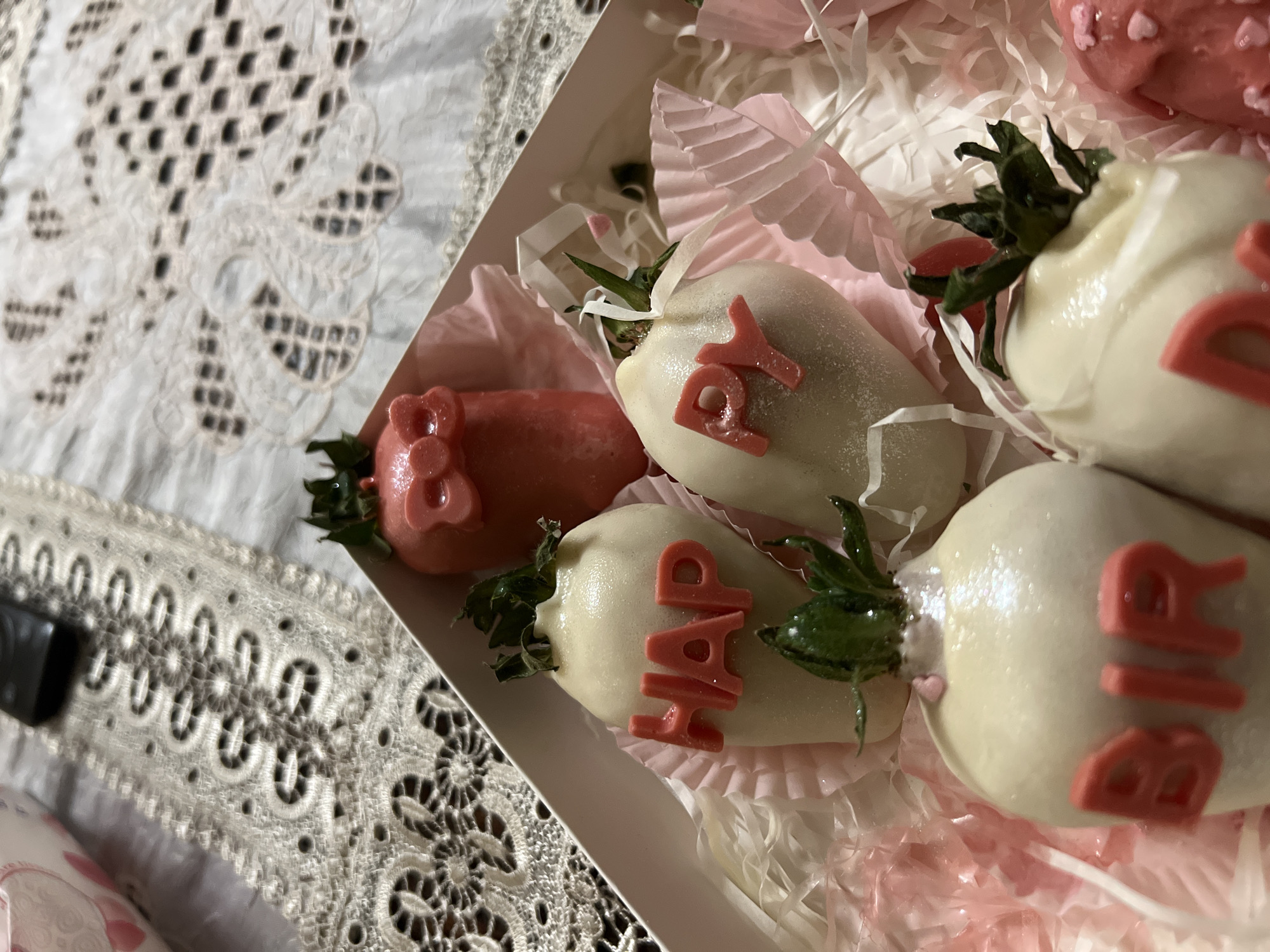 A close-up shot shows a box of chocolate-covered strawberries, some arranged with pink letter decorations that spell out "HAPPY BIRTHDAY." The strawberries are placed on a bed of pink tissue paper. The box sits on a table with a white lace tablecloth. The image location is inferred to be Yerevan, Armenia, based on prior context, but there are no visual indicators in the image itself to confirm this.
