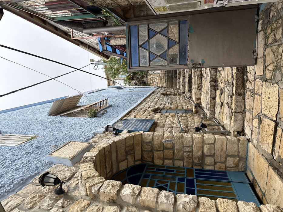 This image depicts a narrow, ascending stone-paved alleyway in Safed, Israel, under an overcast sky.
On the left, a building features a light-colored, rough-hewn stone base that transitions to a textured, light blue stucco upper facade. An arched entrance on the lower left frames a blue metal door with geometric blue and yellow glass-like panels. A black, antique-style wall lantern is mounted on the stone wall above the arch, with a smaller similar lamp lower down. Two rectangular plaques with Hebrew text are affixed to the blue stucco. Higher on the blue wall, a window is partially obscured by a corrugated metal awning, with visible small plants. A silver metal handrail extends along the stone wall, paralleling the ascending steps.
On the right, a multi-paneled metal gate, painted a dull grey with visible chipping, is partially open into the alley. This gate features a large Star of David design composed of blue and clear glass panes. Further back on the right, another building with stone construction and multi-colored window frames (green, red) is partially visible.
The alley floor consists of irregularly shaped, light-colored stone paving. A series of approximately 15 stone steps ascends into the background. Overhead, several black utility wires crisscross the sky. In the distance, at the top of the steps, a light pole and some green foliage with yellow fruit are discernible. No individuals are present in the scene.