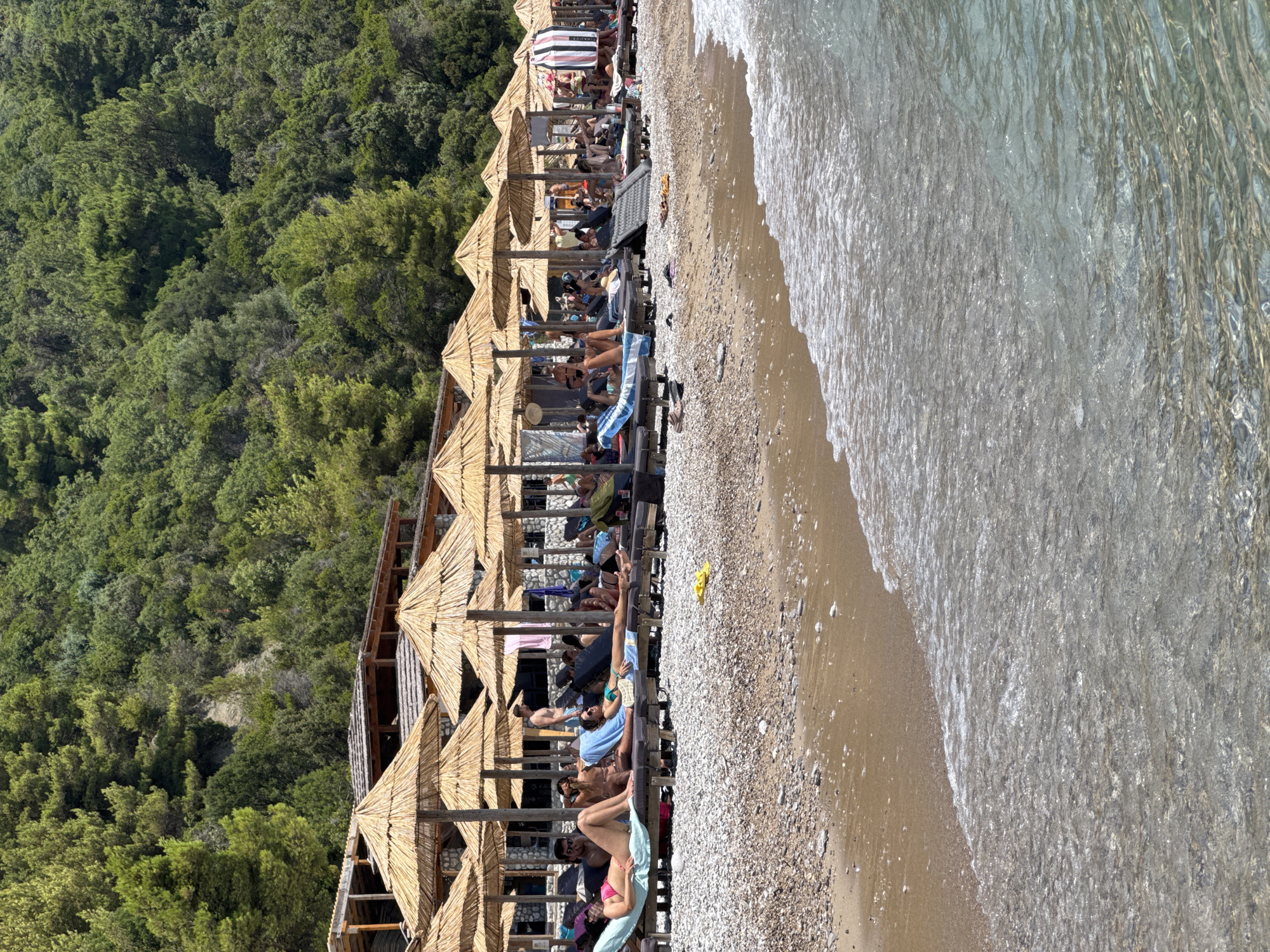 The image shows a crowded beach with many people relaxing on lounge chairs under straw umbrellas. The beach is made of pebbles, and the water is clear and shallow, with gentle waves lapping at the shore. Behind the beach, there is a dense green forest that covers a hillside. There is a wooden structure with a roof behind the umbrellas, which may be a bar or restaurant. Several people are visible on the beach, sunbathing, talking, and some appear to be reading. The overall atmosphere is one of a busy and popular summer vacation spot.