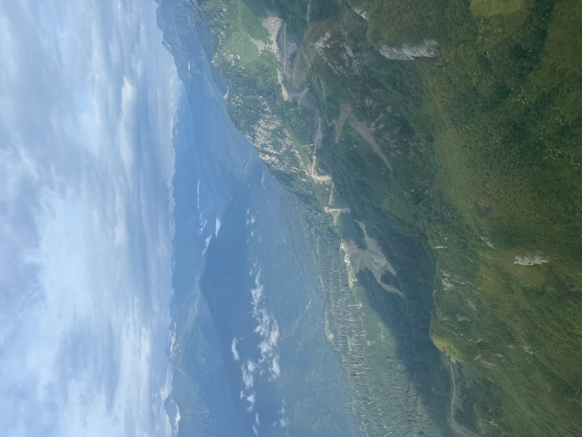 This aerial photograph captures a vast mountain landscape, likely taken from an aircraft, overlooking the area of Estosadok, Russia. The scene is dominated by multiple layers of rolling mountains stretching into the distance, their peaks softened by a hazy, atmospheric perspective. The sky above is filled with a mixture of light and dark clouds, suggesting an overcast or partly cloudy day.
In the foreground and midground, the mountains are densely covered with lush green vegetation, appearing as a thick carpet of trees and undergrowth. Patches of exposed rock and steep slopes are visible, indicating the rugged terrain. Interspersed within the green slopes, there are visible signs of human development, including what appear to be ski slopes or mountain trails winding their way down the mountainsides. Several structures, possibly buildings or ski lift stations, are clustered on some of the higher elevations, identifiable by their white roofs and surrounding infrastructure. Poles and cables, consistent with ski lift systems, are also clearly discernible.
The lighting in the image is diffused, with some areas illuminated by sunlight creating subtle shadows that highlight the contours of the mountains. The overall color palette is dominated by greens and blues, with muted tones due to the atmospheric conditions and distance. No people are visible in the photograph. The environment is natural and mountainous, with clear evidence of its use as a recreational area, likely for skiing or hiking during warmer months.