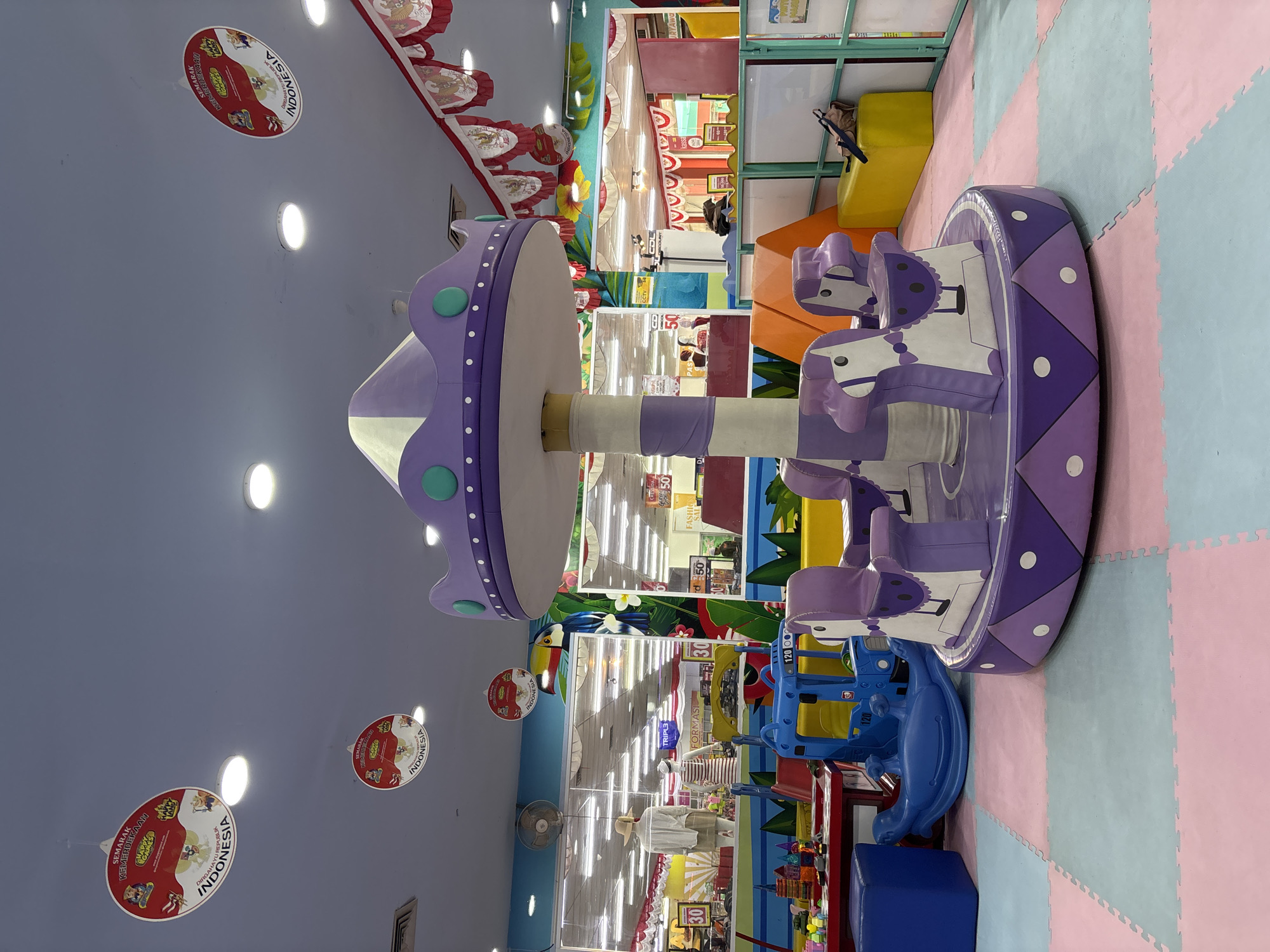 An indoor playground features a purple and white carousel with three attached horse-shaped seats. The carousel is situated on a floor covered with interlocking foam mats in pink, blue, and light green. In the background, there is a blue toy vehicle with "120" printed on its side, and a blue rocking horse. Various other toys, including a red table with building blocks, are visible. The ceiling is a light blue, with several round light fixtures and decorative circular banners hanging down, featuring Indonesian national symbols and text. Red and white festive bunting is also strung across the ceiling. In the far background, through glass partitions, shelves stocked with merchandise and store displays are visible, suggesting this playground is located within a larger commercial establishment, likely a department store or supermarket. The overall impression is a brightly lit, playful environment.