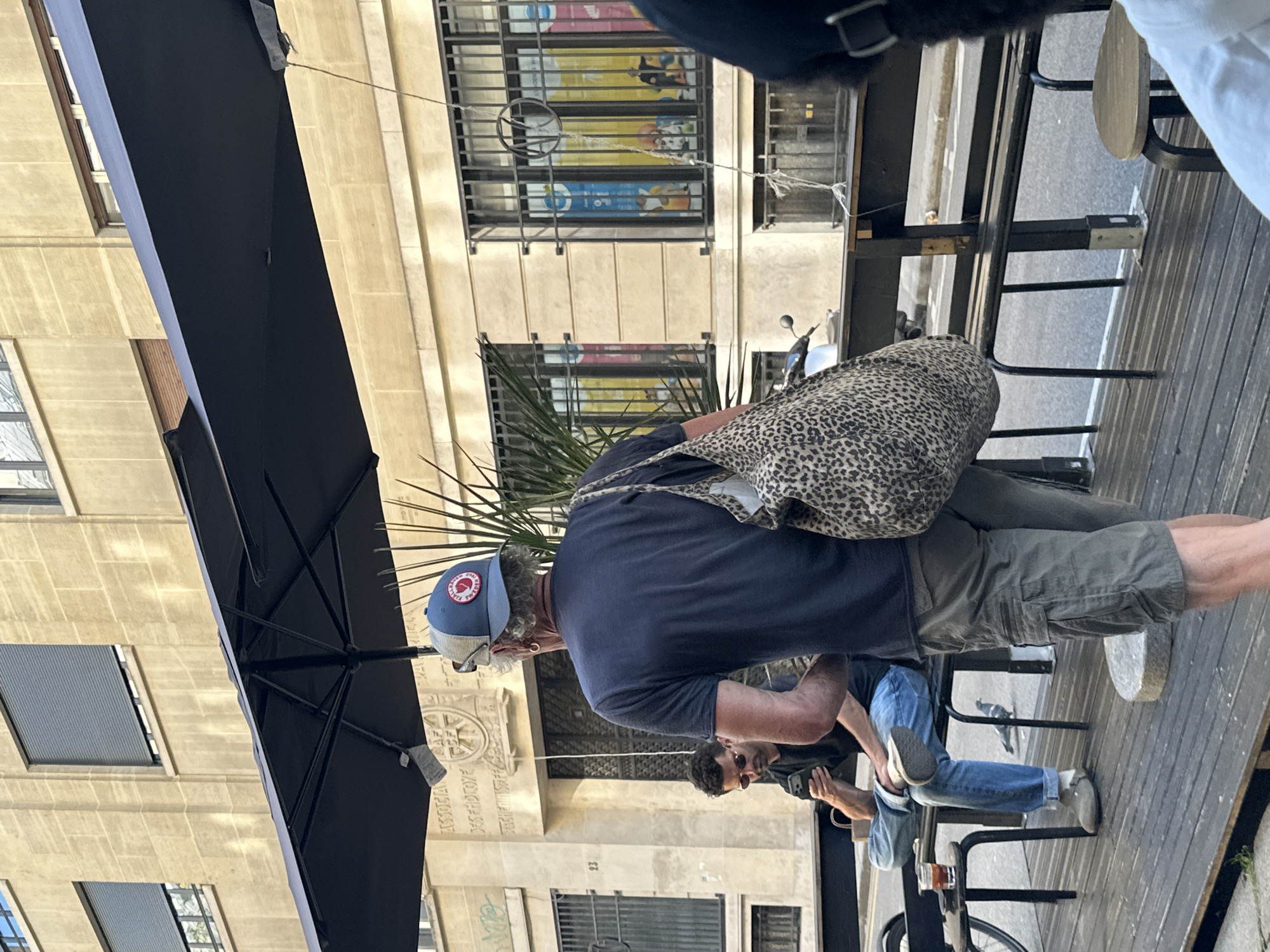 The image depicts a street scene in Paris, France. An older man wearing a blue t-shirt, green shorts, and a blue and white baseball cap with a red logo is standing with a large, spotted bag over his shoulder. He appears to be interacting with another man seated at a table, who is wearing jeans and sneakers and holding a smartphone. A black umbrella is partially visible, providing shade. The setting is an outdoor café or patio, with wooden flooring and metal chairs. In the background, a building facade is visible, featuring multiple windows with metal bars and decorative architectural details. A bicycle is partially seen in the foreground, near the seated man.