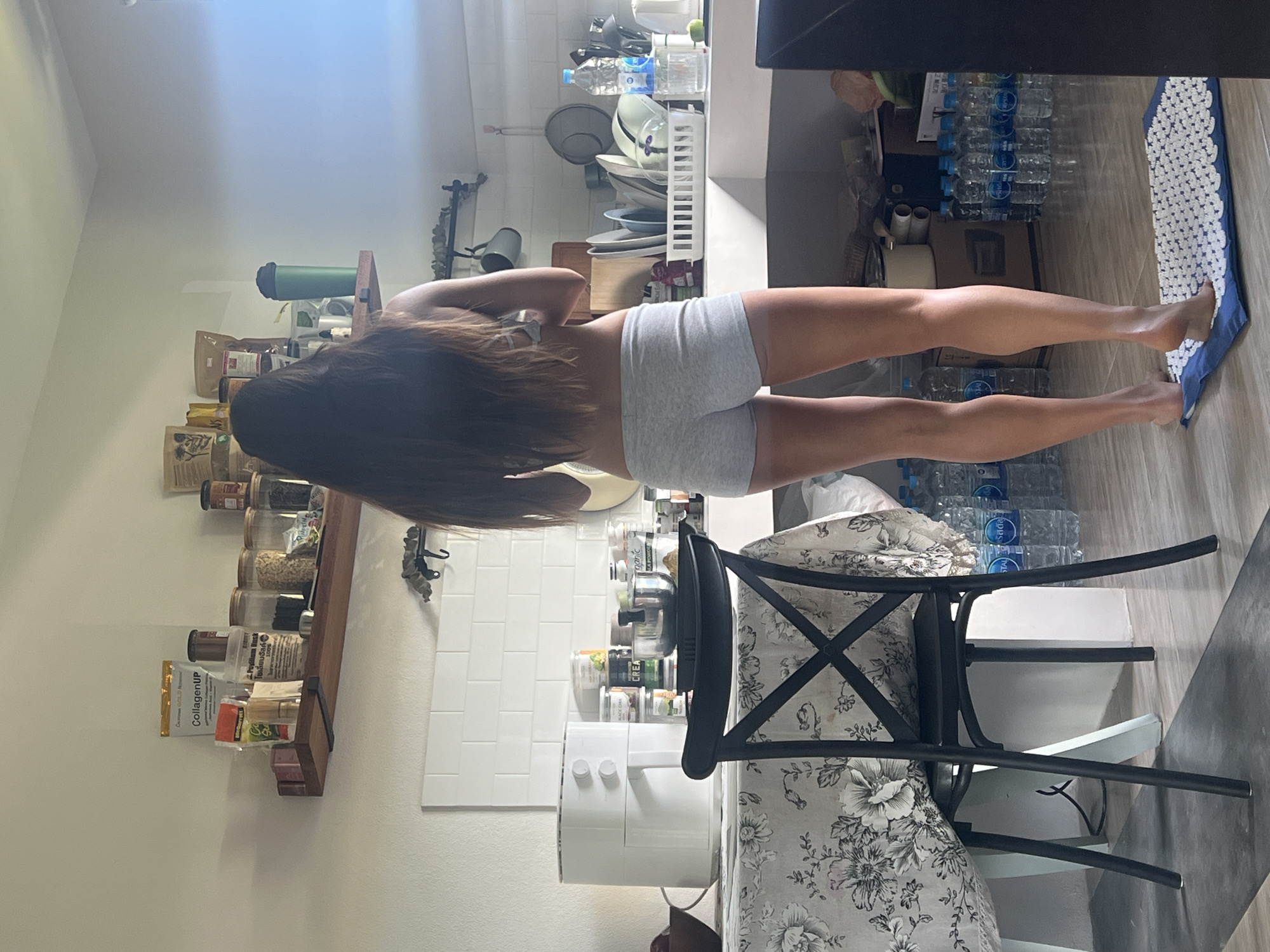 A woman stands in a kitchen in Ban Bang Rak, Thailand. She is seen from the back, wearing grey shorts. Her dark hair falls past her shoulders. The kitchen has white tiled walls and a wooden shelf above the counter, displaying various jars and containers. A white air fryer is on a table covered with a floral cloth, beside a black chair. Water bottles are stacked on the floor to the right of the woman, and she is standing on a blue and white mat. The room is brightly lit by natural light.