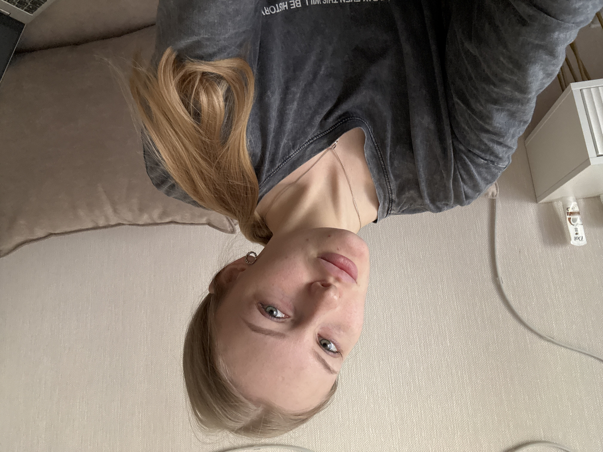 A woman with fair skin, light blue eyes, and blonde hair, part of which is styled in a low ponytail, is pictured from a slightly downward angle. She is wearing a grey, v-neck t-shirt and a delicate silver necklace. The background includes a light beige cushion of a sofa or couch in the upper left and a light beige wall with a white shelf unit and some product bottles in the upper right. It's difficult to pinpoint the precise location in Perm, Russia, but the environment appears to be an indoor setting, possibly a home or apartment, based on the furniture and décor. The photograph is rotated, with the woman appearing upside down in the frame.
