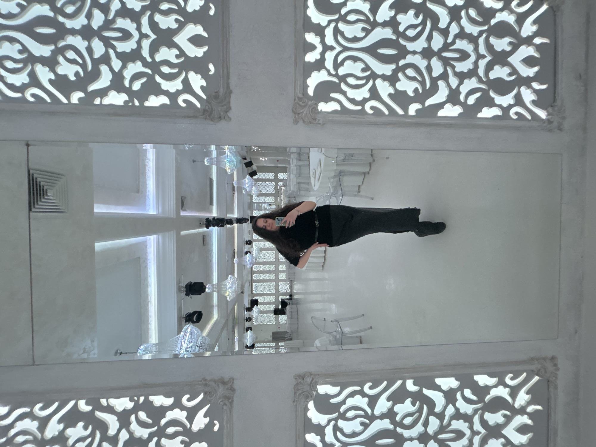 A woman wearing black clothing is taking a picture of herself in a large, vertical mirror. The mirror reflects the interior of a room, including ornate chandeliers, white chairs, and decorative wall panels with white, floral-like cutouts. The room appears to be a formal event space. The location may be in Plovdiv, Bulgaria.