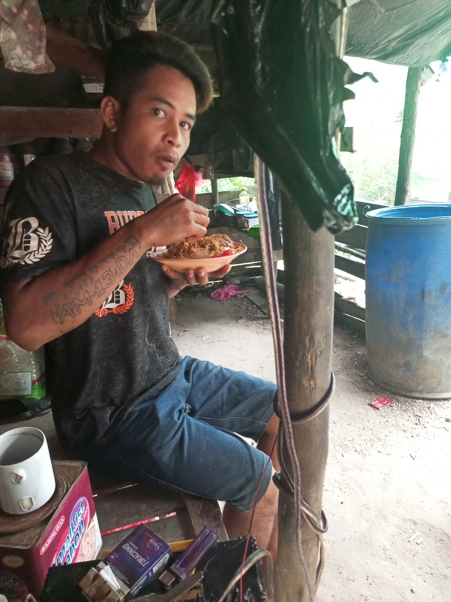 An Indonesian man, identified by a tattoo on his forearm reading "AM-13 DA," is seen eating from a plate of noodles while sitting outdoors in what appears to be a makeshift shelter. The shelter's structure consists of wooden posts and a plastic tarp overhead. In the background, a large blue plastic barrel and various other items, including a white mug, a box of cigarettes, and a lighter, are visible. The ground is covered with gravel and dirt. The overall scene suggests a simple, possibly rural, living environment within the city of Keling Panggau, Indonesia.