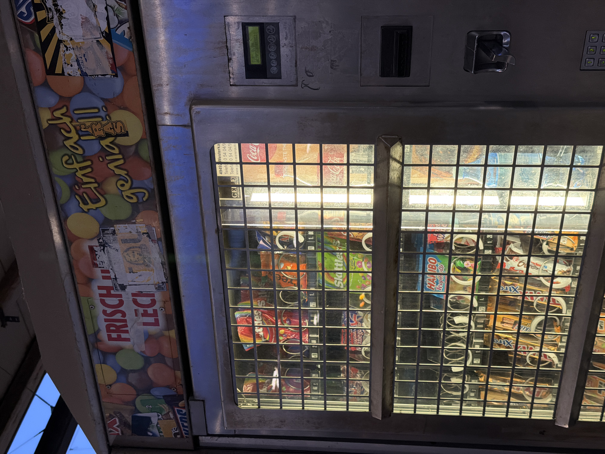 The image shows a close-up view of a vending machine that appears to be located in Witten, Germany. The machine is filled with various snacks and drinks, including bags of Skittles, Haribo Tropifrutti, and Coca-Cola bottles. The top of the machine features a colorful advertisement with German text that reads "Frisch & Leck" and "Einfach genial!". The machine itself is made of metal and has a glass display window showing the products inside. A digital display on the right side of the machine shows "EUR 0,00" and some German text. The overall environment suggests an outdoor setting, possibly a street or a public area, given the lighting and the slightly weathered appearance of the machine and its surroundings. There are no people visible in the image.
