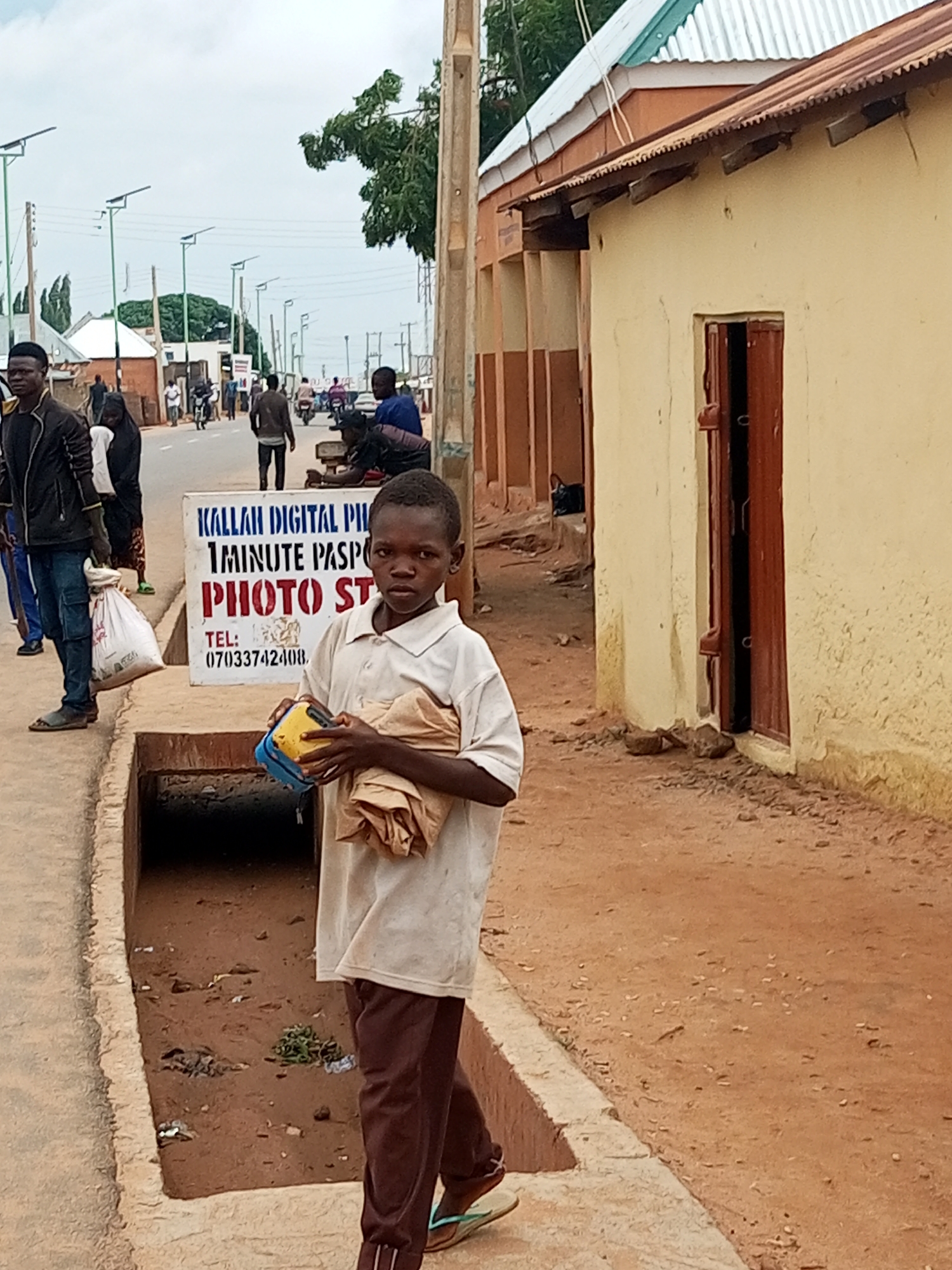 In Tempure, Nigeria, a young boy stands on a dirt path next to a concrete drainage ditch. He is holding a yellow and blue device and a bundle of tan fabric. He wears a light-colored polo shirt and brown pants. Behind him, a sign advertises "KALLAH DIGITAL PHOTO STUDIO" and provides a phone number. A group of people and motorcycles are visible on a road in the background, with solar-powered streetlights lining the street. The scene is set against the backdrop of a building with a corrugated metal roof.