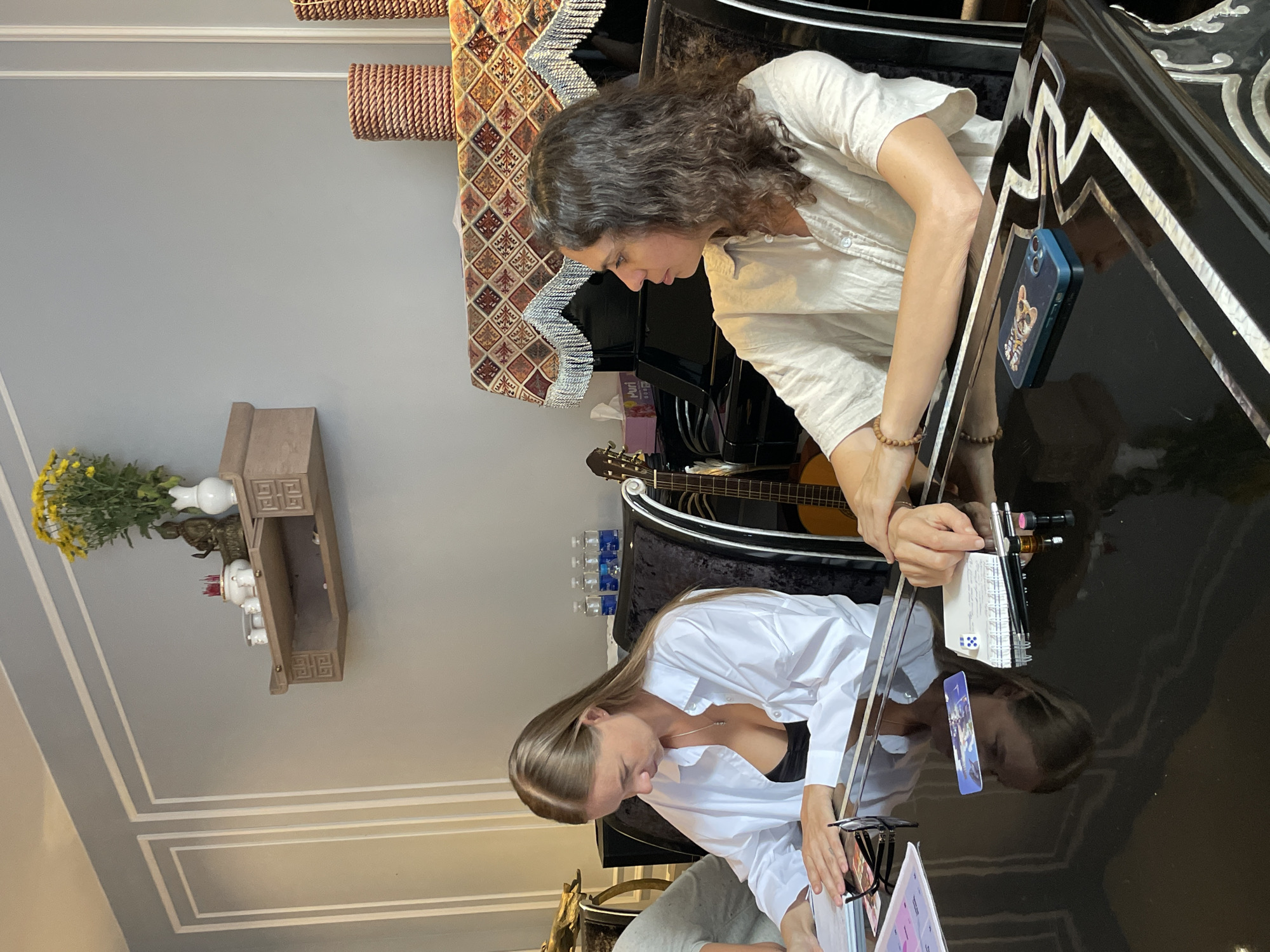 Two women are gathered around a glossy black table, which reflects their images. The woman in the foreground, with long blonde hair, wears a white open-necked shirt over a black top and is looking down at a notebook or papers. The woman behind her, with curly brown hair, is wearing a light-colored linen shirt and is also looking down, with one hand resting on the table. There are various objects on the table, including a phone with a cat case, pens, a notebook, a small die, and a guitar is visible behind the women. In the background, a shelf is mounted on a light grey wall, holding a vase with yellow flowers, a small statue, and what appear to be incense sticks. The overall scene suggests a moment of study, work, or shared activity. The lighting appears to be indoor ambient light. The location is identified as Xuân Trường - Đà Lạt, Vietnam.