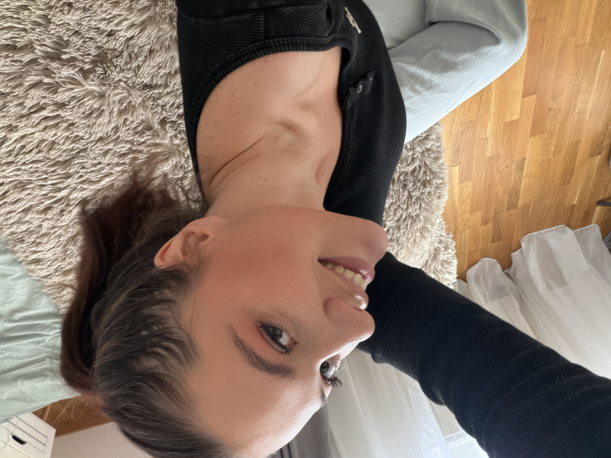 A woman is lying on a bed or floor covered with a beige shag rug, wearing a black long-sleeved top and light blue or grey pants. Her head is positioned at the bottom left of the frame, and she is looking towards the camera. The background includes light-colored curtains near a window and a hardwood floor. The scene's location is not discernible based on the provided image. There is no indication the scene is in Belgrade, Serbia.