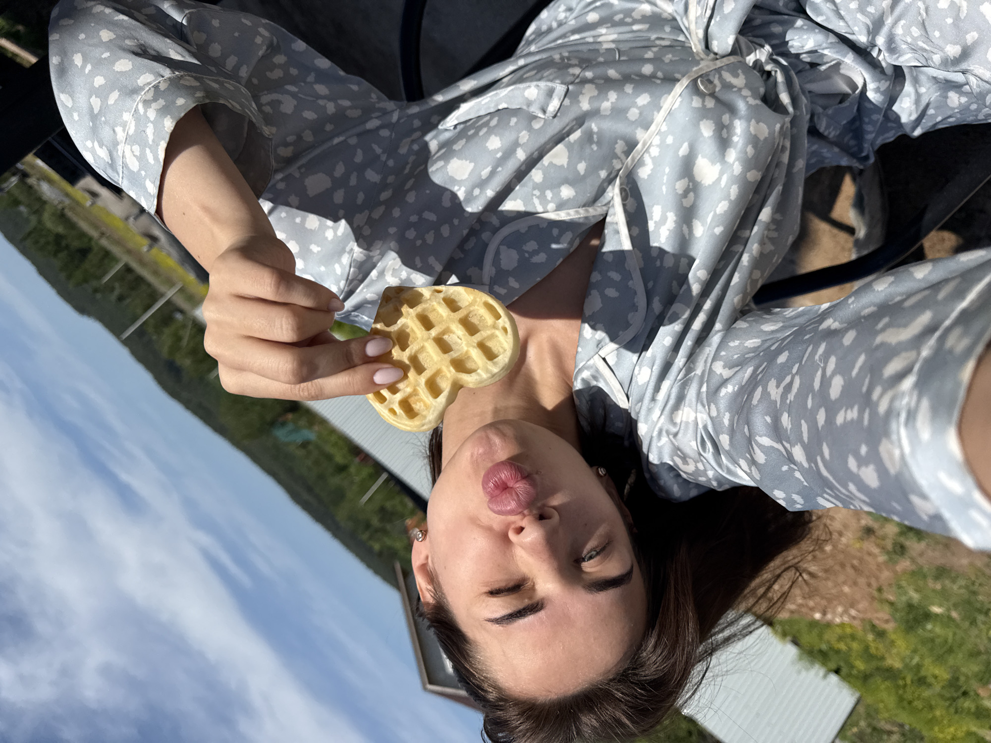 A woman with brown hair is taking a selfie outdoors, holding a heart-shaped waffle in her right hand. She is wearing a light blue patterned pajama set and has her lips pursed in a kissing expression. The background shows a partially obscured landscape, suggesting a rural or suburban setting, with glimpses of trees and a light blue sky. Based on the prompt information, the location is Лобаново, Russia.