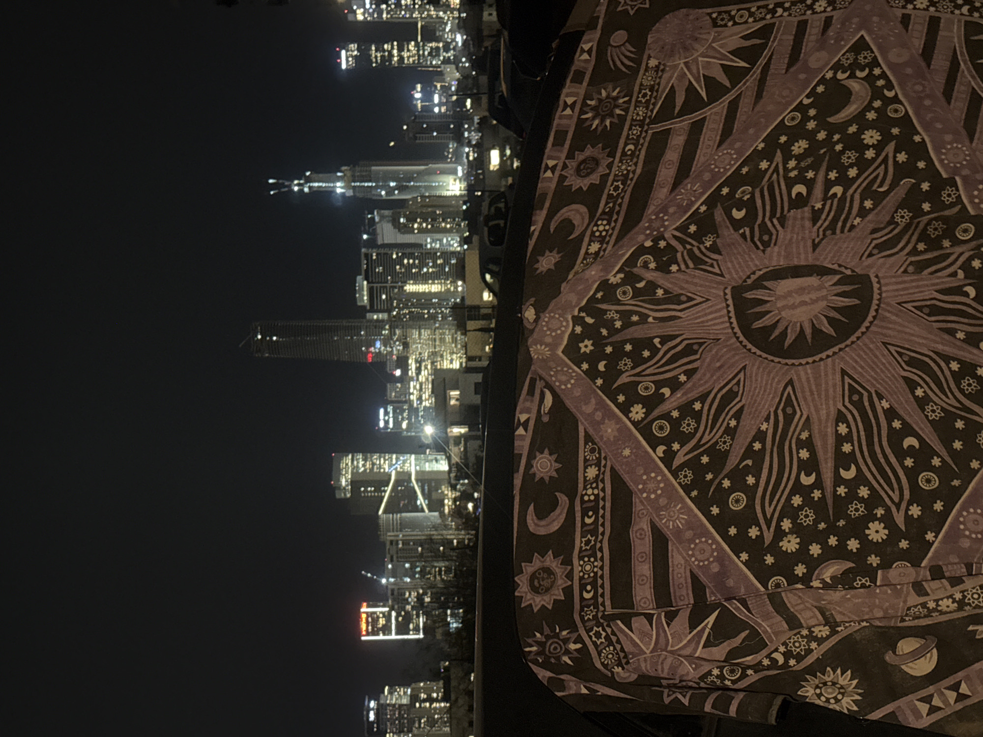 A nighttime view of the Givatayim, Israel skyline, shot from a high vantage point. The skyline consists of high-rise buildings with lighted windows and illuminated tops, under a dark sky. In the foreground, a tapestry-like textile with celestial motifs (suns, moons, stars) is draped over an unseen surface. The tapestry is predominantly dark with light purple designs. A black object is partially visible between the tapestry and the skyline.