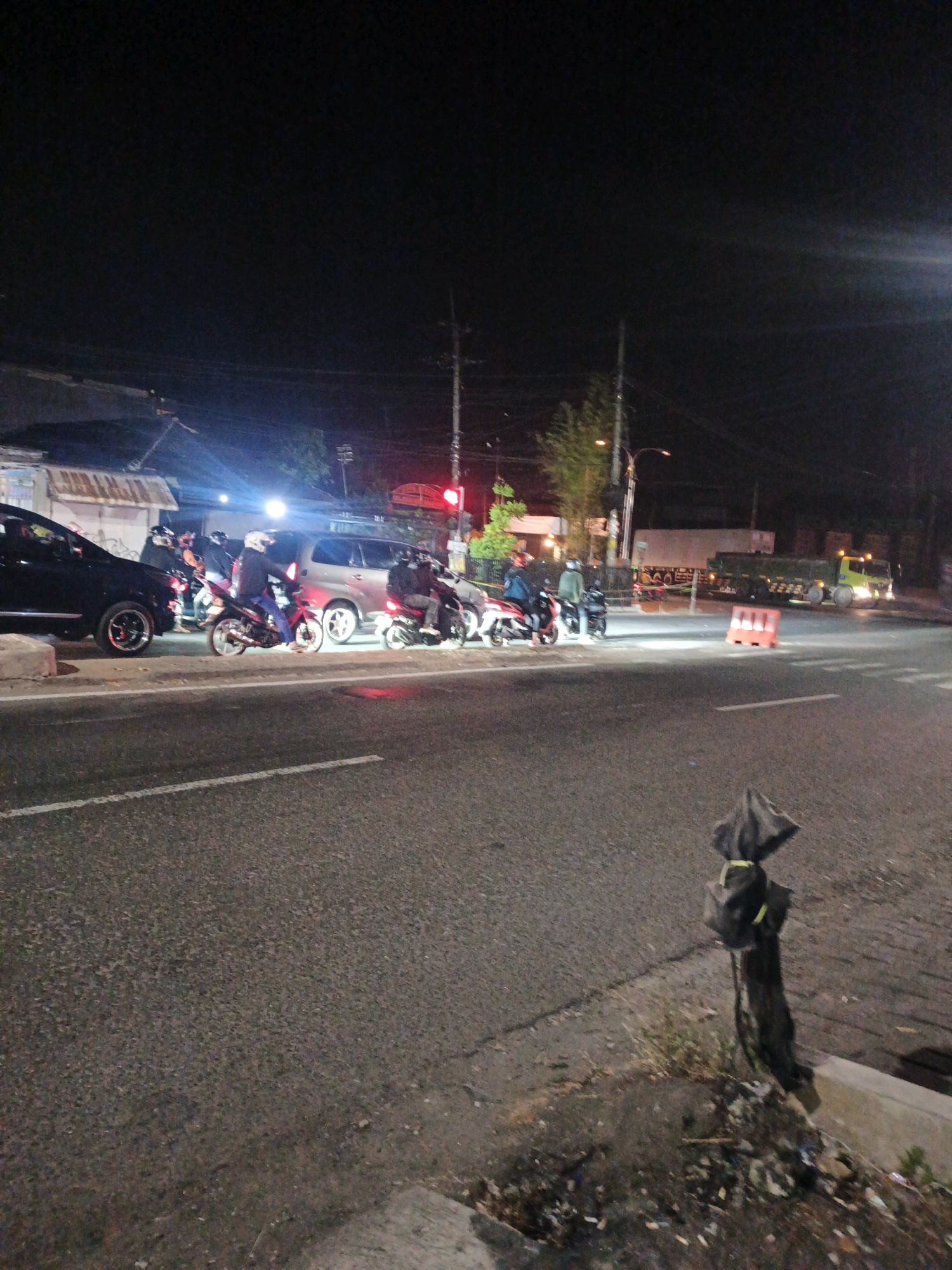 This image depicts a street scene at night in Meri, Indonesia. A black car is parked on the left side of the road, with its headlights on. Several people on motorcycles are gathered near the car, some wearing helmets. A silver minivan is also visible behind the motorcycles. The road is paved, with white lane markings. In the background, there are buildings, trees, and streetlights. A large truck is seen further down the road. On the right side of the image, a utility pole is present, and a black bag is tied to a post. Debris is scattered along the side of the road. The lighting suggests it is a well-lit urban or semi-urban area at night.