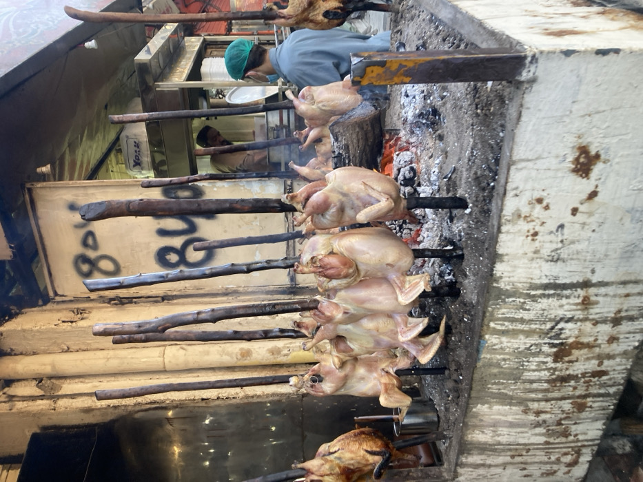 This image captures a vibrant street food scene featuring an open-air chicken roasting setup. In the foreground, numerous whole chickens are being roasted over an open fire, skewered horizontally on thick wooden poles or metal rods. Six chickens are clearly visible, with one at the bottom-most part of the frame appearing significantly more cooked and golden brown, while the others are lighter in color, suggesting they are in earlier stages of cooking. Another partially visible, darker, roasted animal (possibly a small lamb or goat) is at the top left of the roasting line.
Beneath the chickens, glowing red charcoal and embers provide the heat, with some smoke rising. The roasting mechanism appears rustic, built into a weathered, concrete or stone structure that forms the side of the cooking area. Metal bars or supports hold the spits in place over the heat source.
In the mid-ground, behind the roasting chickens, two individuals are visible. One person, wearing a light blue shirt or jacket and a green surgical cap or hairnet, is looking away from the camera, seemingly focused on tasks within an indoor or semi-enclosed kitchen area. Their face is partially obscured by what appears to be a mask. Further back and slightly lower, another person is partially visible, also engaged in work.
The background reveals a simple setting with light-colored walls or panels. On one of these surfaces, large black graffiti-style numbers "8008" are prominently displayed. A white rectangular object with a "York" logo, possibly a refrigerator or cooler, is also visible in the background, along with stacks of white plates or containers. The overall impression is one of a busy, functional, and traditional outdoor cooking environment, likely associated with a local eatery or street vendor. The lighting suggests it could be daytime or a well-lit evening.
