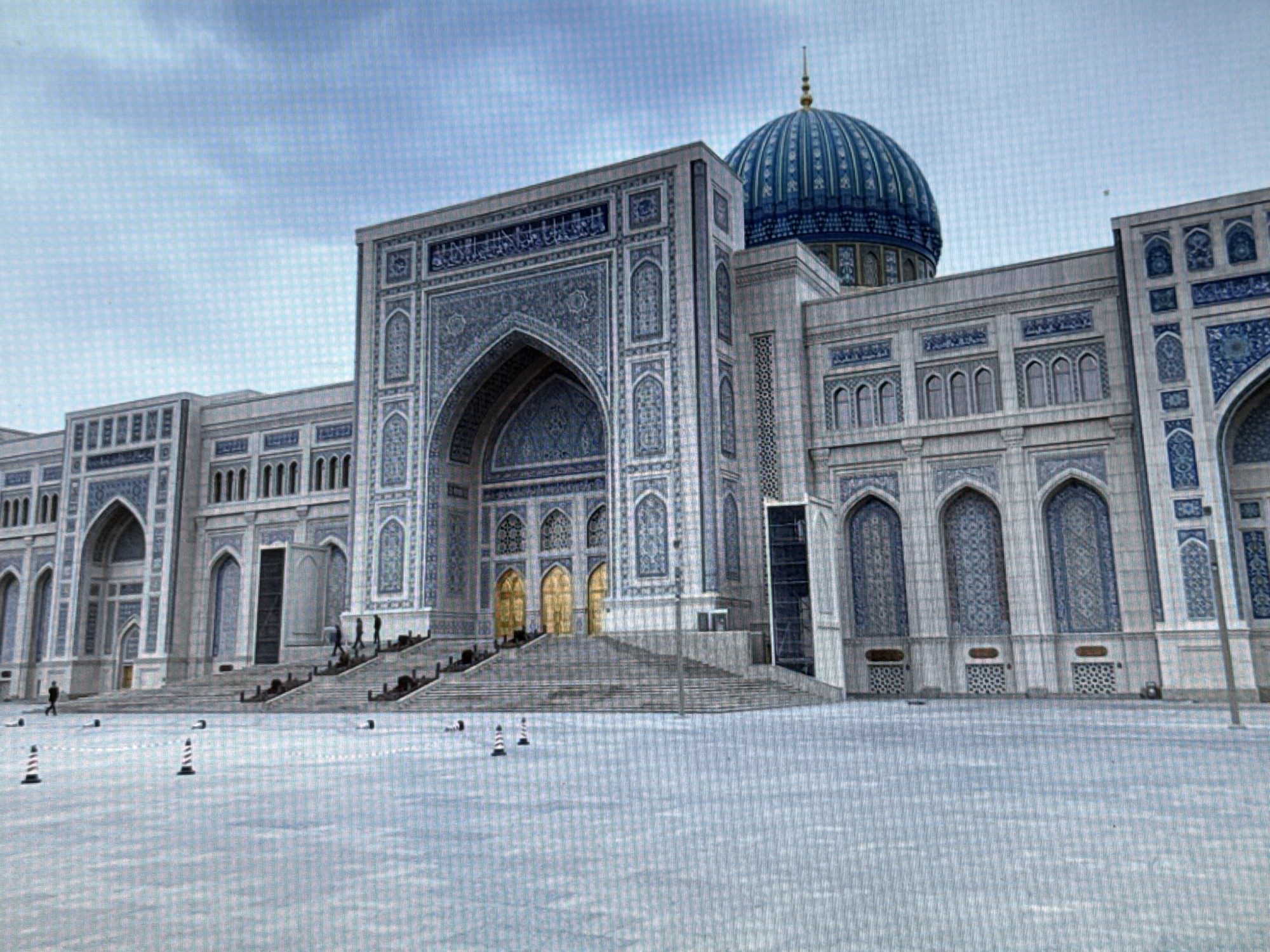 A large, ornate building with intricate blue and white tilework, and a prominent blue dome, is situated in what appears to be a public square. The architecture suggests a religious or governmental structure, possibly a mosque or a palace. The foreground is a flat, open area covered in a light-colored, textured surface, possibly snow or a specially designed pavement. Several traffic cones, some red and white striped and others solid white, are scattered across this area. A few individuals are visible, some walking up a wide staircase leading to the building's entrance, while others are present in the square. The sky is cloudy with visible blue patches. The image is captured from a slightly low angle, emphasizing the scale of the building. The scene appears to be in Pushkino, Russia, based on the provided context.