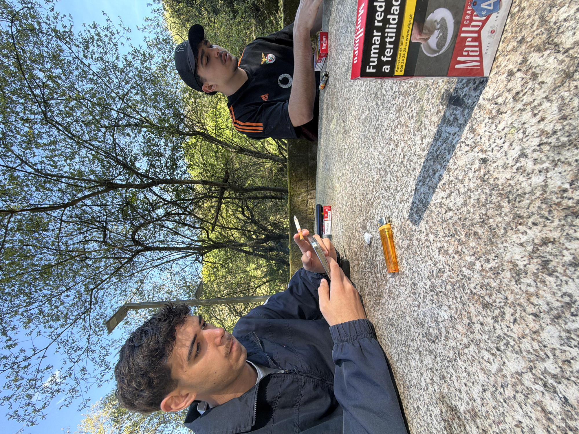 Two young men are sitting at a textured gray stone table outdoors on a sunny day in Vila Real, Portugal. One man, facing the camera, is wearing a dark blue jacket over a white shirt and is holding a lit cigarette in one hand and a mobile phone in the other. The second man, facing away from the camera, wears a black Adidas soccer jersey with orange stripes, and a black baseball cap. On the table are cigarettes, a lighter, and a Marlboro cigarette pack with text written in Portuguese, "Fumar reduz a fertilidade." The surrounding environment includes trees and a clear blue sky, suggesting the location is likely a park or a public space.
