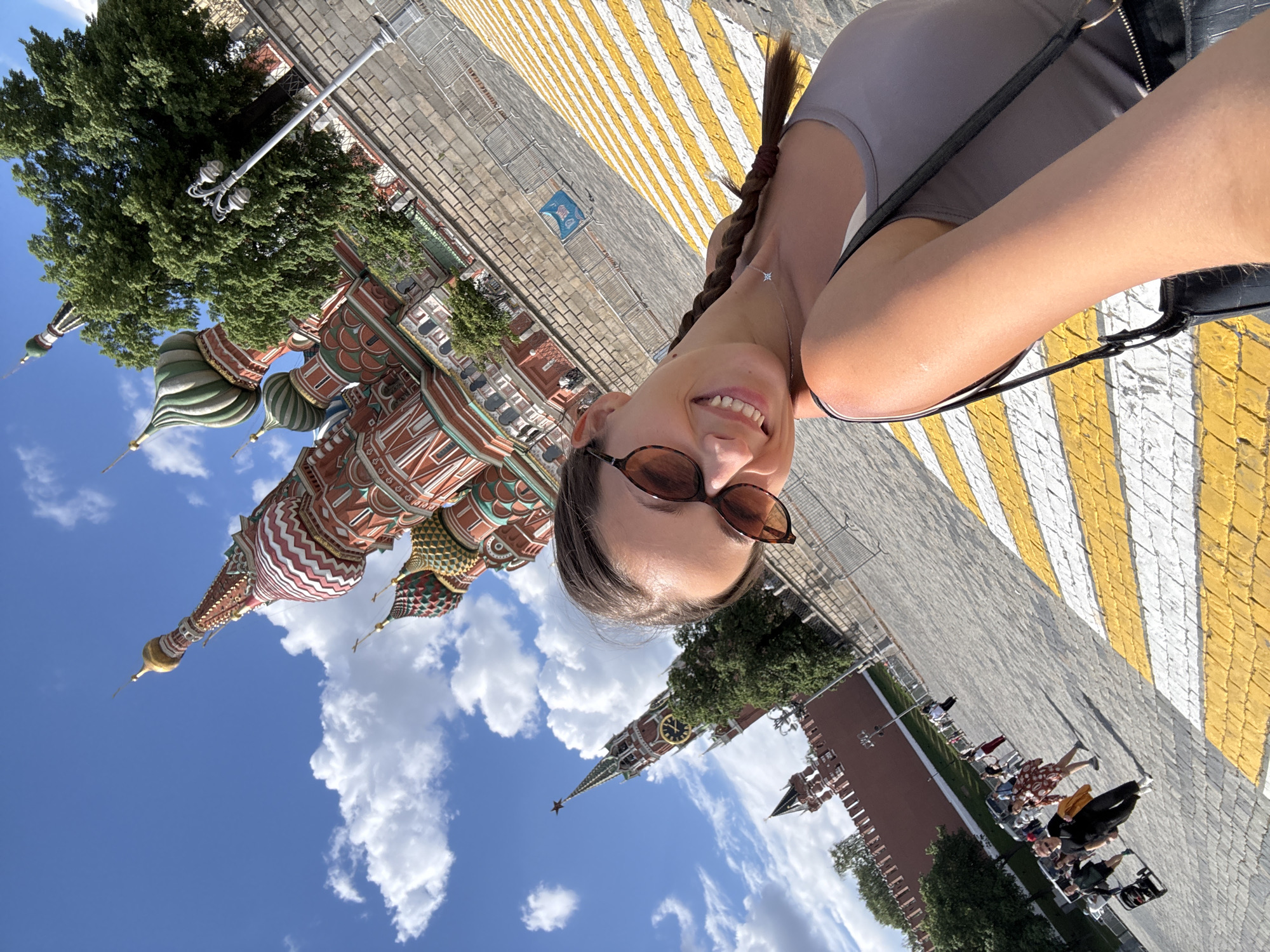 A rotated, eye-level selfie captures a woman with braided hair, sunglasses, and a grey top smiling toward the camera. Behind her is St. Basil's Cathedral with its iconic onion-shaped domes and spires. Blue skies with scattered clouds fill the upper left quadrant. The Spasskaya Tower and segments of the Kremlin wall are visible in the bottom portion of the image. The scene takes place on Red Square in Moscow, Russia. In the lower right quadrant, a pedestrian crosswalk with yellow and white stripes and a row of people in wheelchairs are visible.