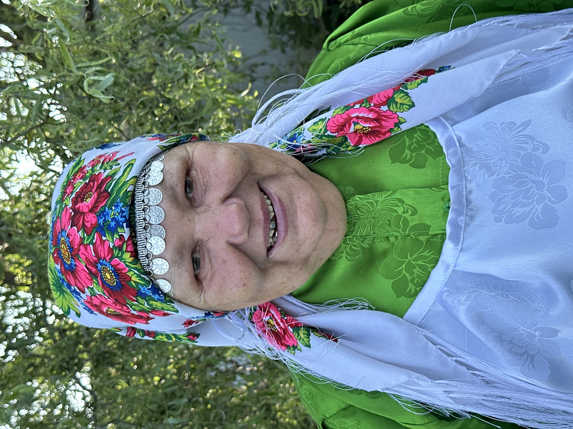 A middle-aged woman is pictured outdoors, with a blurred green foliage background suggesting a natural or garden setting, possibly in the village of Sredneye Kechevo, Russia. She is wearing a traditional outfit consisting of a brightly colored floral headscarf, a green patterned blouse, and a white embroidered apron with fringe detailing. The woman is smiling, revealing some dental work, and is looking directly at the camera. The lighting suggests it is daytime.
