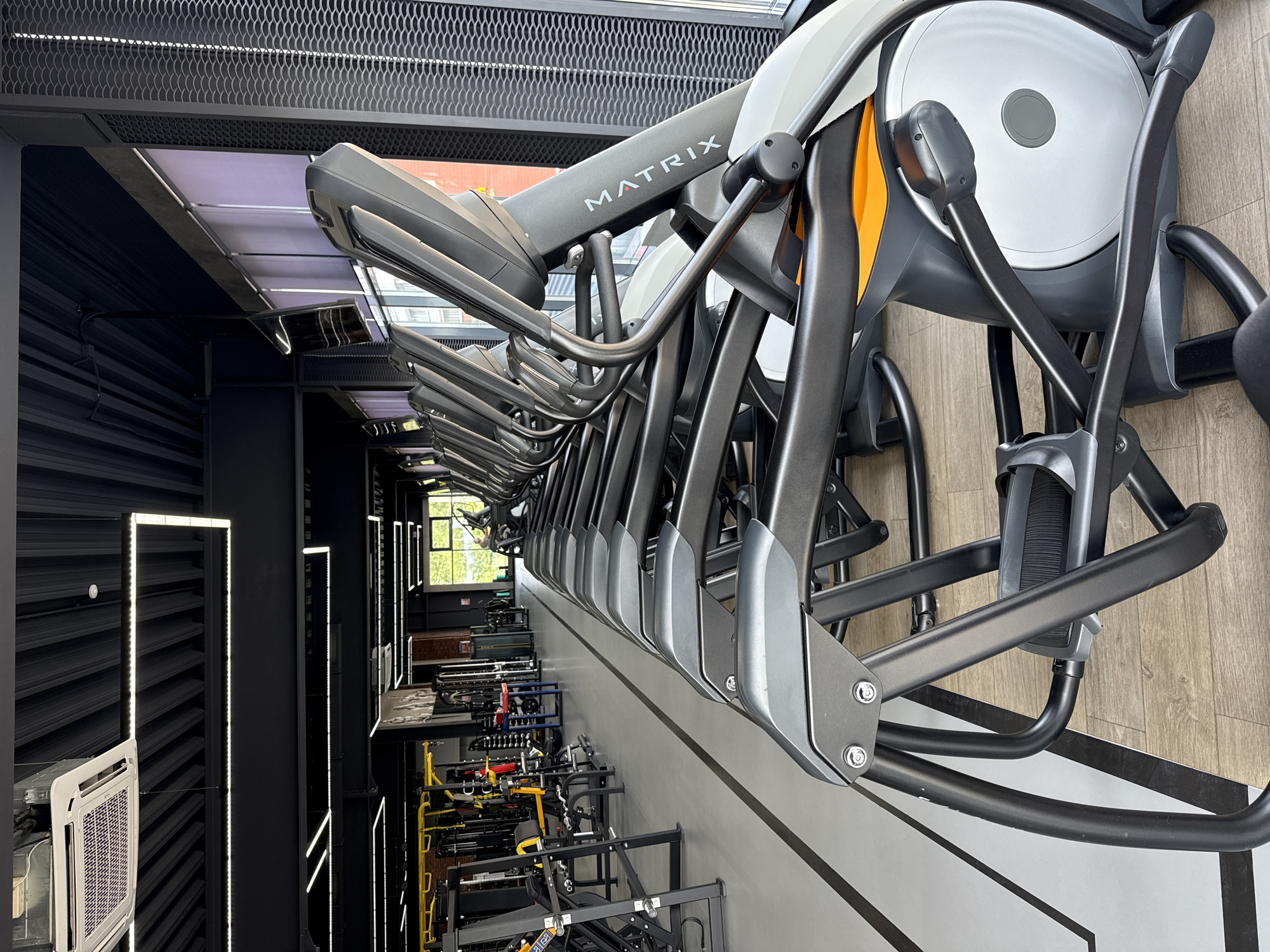 The image shows a row of Matrix-branded elliptical machines in a gym. The machines are dark gray and black with some orange accents. The gym has a modern industrial aesthetic, with black metalwork, exposed ductwork, and LED strip lighting. In the background, beyond the ellipticals, is weight training equipment and a window. The specific location within Votkinsk, Russia, is not discernible from the image. The image primarily shows exercise equipment in an indoor gym environment.