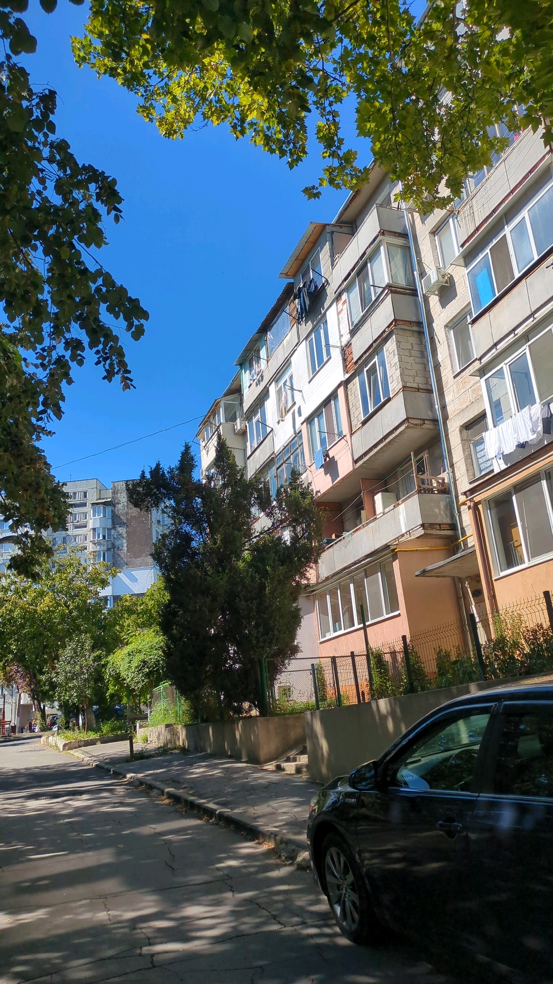 The image shows a street scene in Chișinău, Moldova, on a sunny day. In the foreground, a black car is parked on the side of the road. The road is paved with asphalt and has visible cracks. A sidewalk runs alongside the road, bordered by a low concrete wall and a metal fence. Behind the fence, there is a building with multiple balconies, some of which have laundry hanging out to dry. Green trees and foliage are visible throughout the scene, with branches from a tree in the upper left corner partially obscuring the clear blue sky. In the background, other residential buildings can be seen. The overall impression is of a typical urban environment.