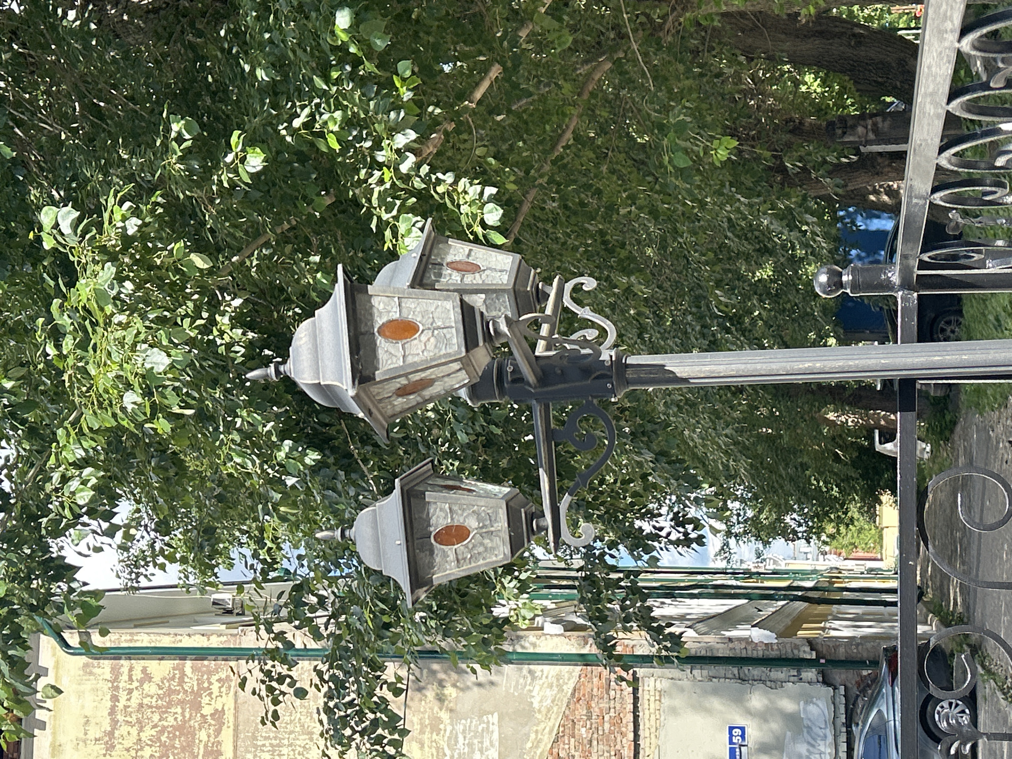 The image shows a decorative street lamp with three heads, set against a backdrop of lush green trees and buildings. The street lamp is made of dark metal and features ornate scrollwork. Each lamp head is enclosed in glass panels, with an amber-colored accent in the center of each panel. The building in the background appears to be old, with a textured facade that shows signs of age and wear. A street sign with the number "59" is visible on the building. A black metal fence is in the foreground. The scene is bathed in sunlight, casting shadows and highlighting the details of the lamp and surrounding foliage. This photograph was taken in Perm, Russia.