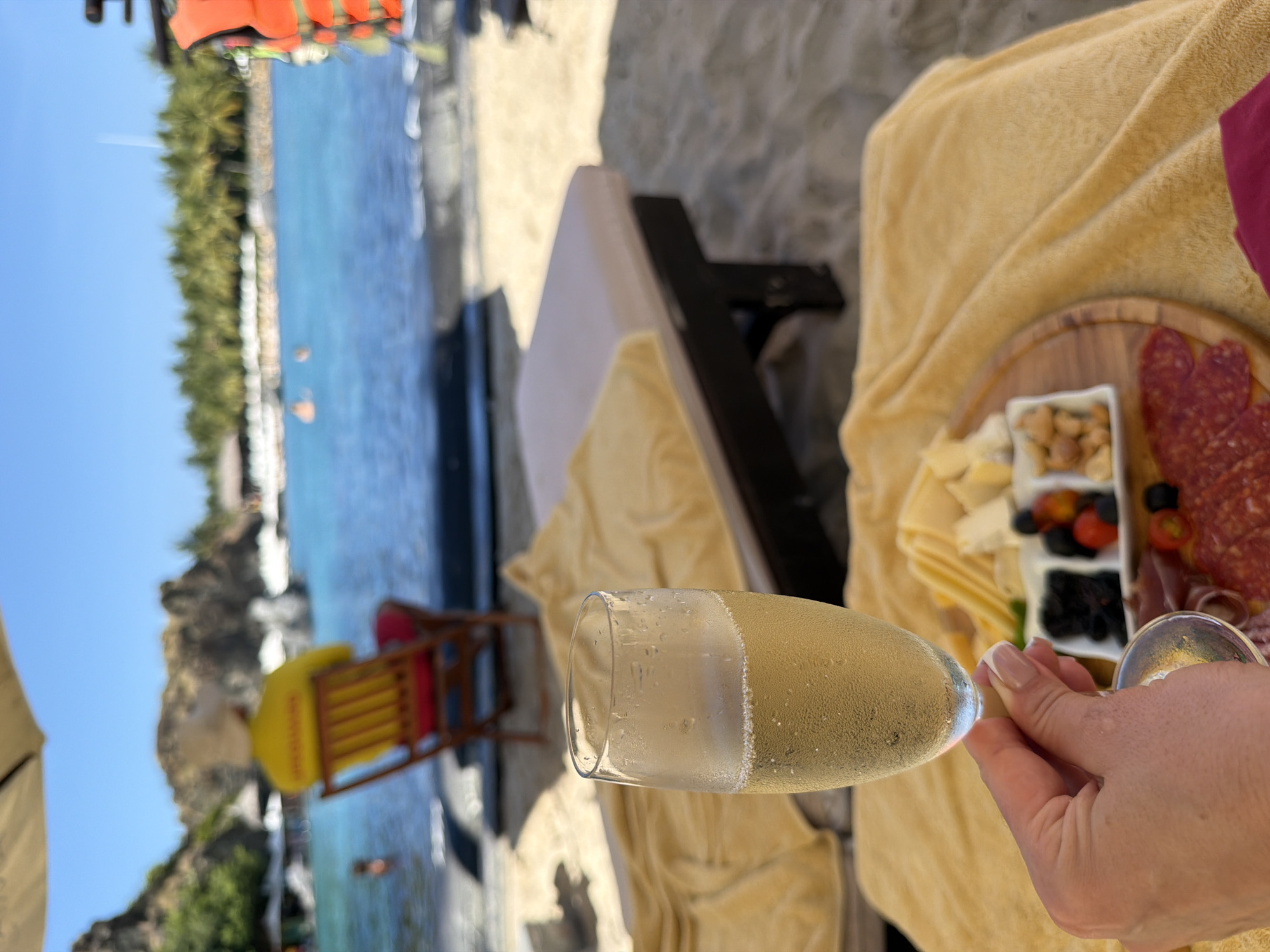 The image, taken in Bac Nha Trang Ward, Vietnam, shows a beach setting. In the foreground, a hand holds a glass of champagne, with a platter of cheese, nuts, olives, tomatoes, and salami adjacent. A yellow blanket covers a beach chair. Further back, there is a red and yellow lifeguard chair on the beach. The beach is sand and leads to blue ocean water. Trees and possibly cliffs are visible on the horizon. The sky is clear blue.