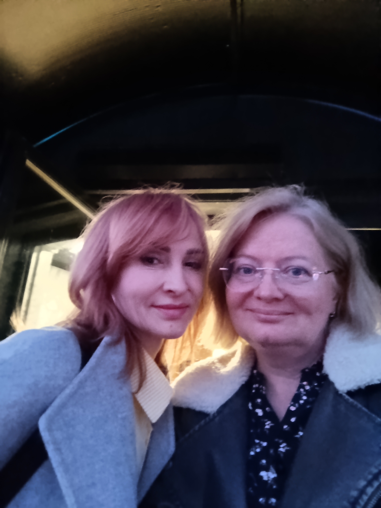 A close-up portrait features two women standing side-by-side, likely taken indoors or in a covered setting in Chaikovsky, Russia. The woman on the left has reddish-blonde, shoulder-length hair with bangs, and a light complexion, wearing a light gray coat over a pale yellow collared shirt. She displays a gentle smile. Adjacent to her, the second woman, appearing older, has blonde hair and wears clear-framed glasses. She is dressed in a dark jacket with a prominent cream-colored, fluffy collar, and a dark blouse featuring a light floral pattern. She also exhibits a soft, pleasant smile.
The background is largely dark and indistinct, suggesting an enclosed or heavily shaded area, possibly a tunnel, underpass, or the interior of a large structure. Dark structural elements, such as beams or an arch, are faintly visible in the upper portion of the frame. Light illuminates the subjects primarily from the left, casting a warm tone on their faces and hair. Both women appear relaxed and content, sharing a pleasant moment captured in the photograph. No text is visible in the image.