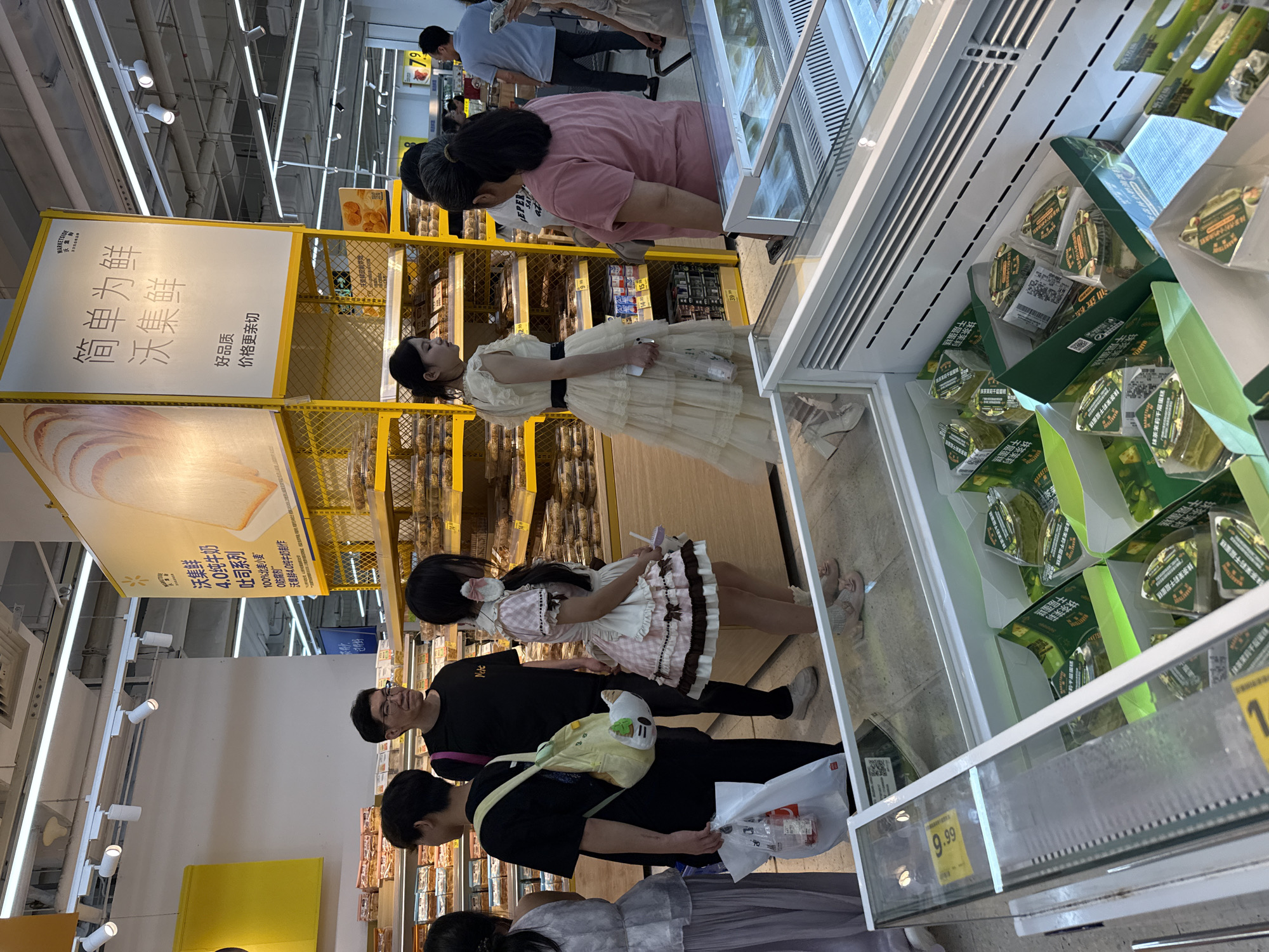 The image shows a well-lit interior of a supermarket, possibly a Walmart, in Huizhou, China. In the foreground, a refrigerated display case contains numerous green packaged items, appearing to be some kind of food product, possibly desserts or pastries. The products are arranged in boxes and individual servings, with Chinese text visible on the packaging.
In the mid-ground, several people are browsing the aisles. On the left, a person in a light-colored dress is partially visible. Next to them, a person in a black t-shirt and black pants holds a white plastic bag with a bottle inside and carries a yellow backpack adorned with a cartoon character. Behind them, a man wearing glasses and a black t-shirt stands looking forward. To the right of the man, a young woman in a pink and white checkered dress with a white apron stands with her head turned. Further to the right, a woman in a cream-colored tiered dress with a black belt is speaking to another woman in a pink t-shirt. In the background, more shelves are stocked with food items, and other shoppers can be seen. A large yellow sign with Chinese text and images of bread is prominently displayed on a shelf unit. The overall environment suggests a typical retail setting.