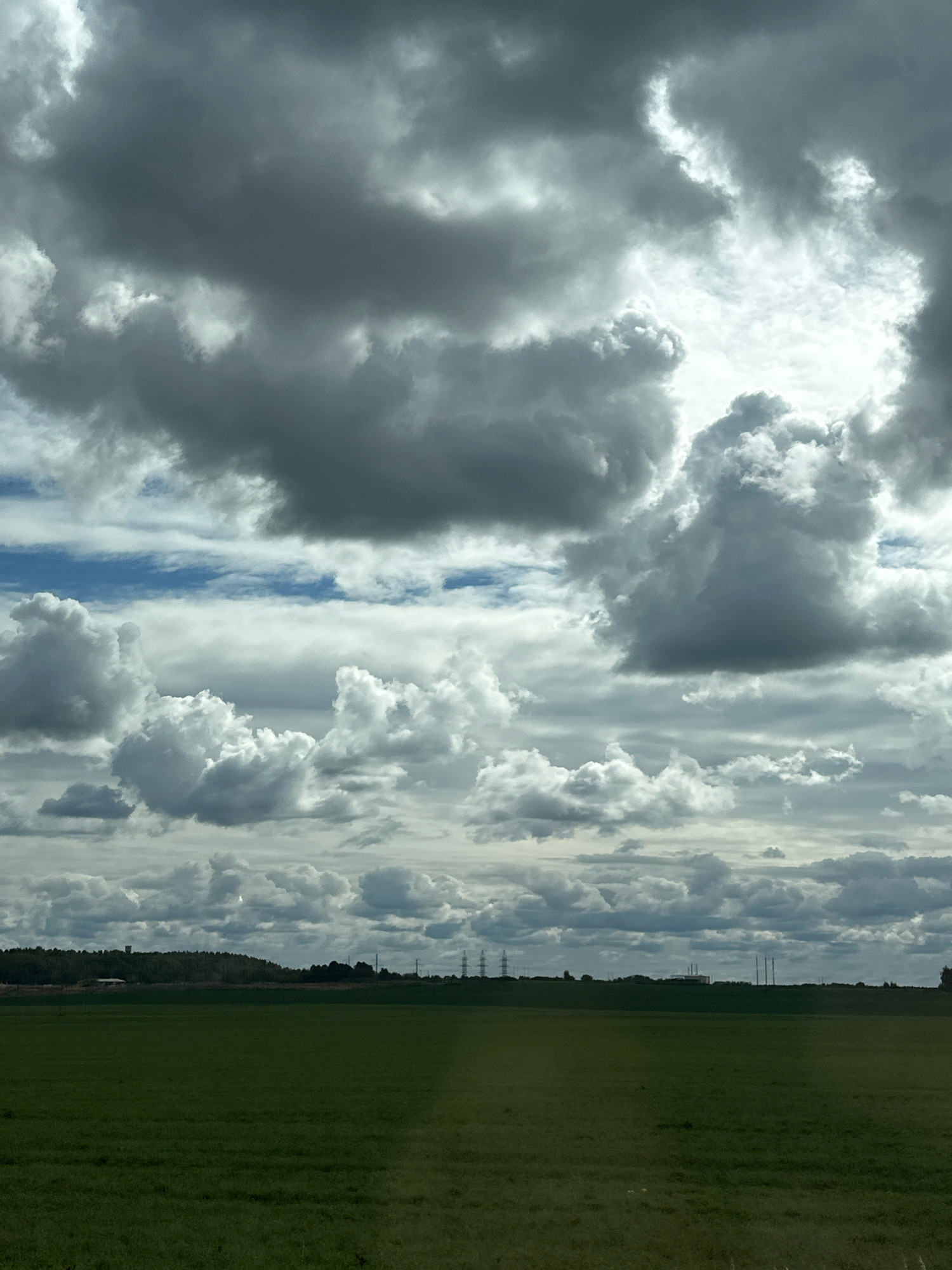 The image depicts a rural landscape under a dramatic sky filled with a mix of dark, heavy clouds and brighter, billowy formations. The foreground consists of a vast, green, grassy field. A line of trees and vegetation forms a distant horizon, beyond which several tall, industrial-looking structures, possibly power line pylons or communication towers, are visible. A few low-lying buildings are also discernible against the tree line. The overall impression is one of a wide-open, somewhat overcast, agricultural or undeveloped area within the territory of Блонскі сельскі Савет, Belarus. There are no people or animals visible in the frame.