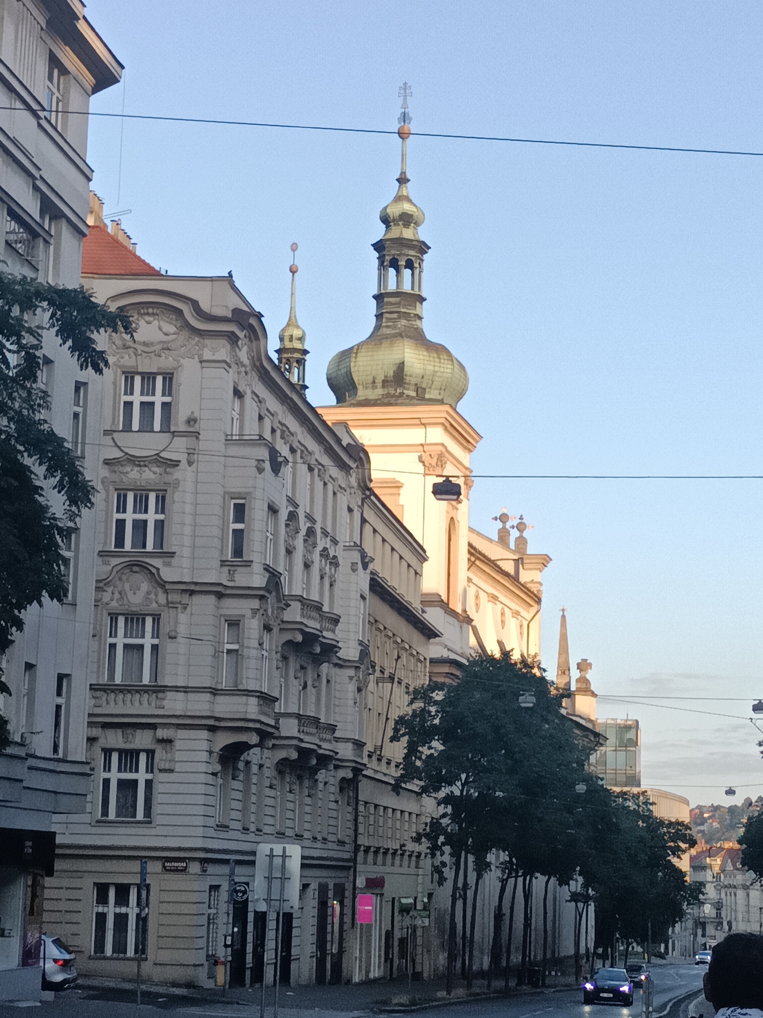 This is a vertical, eye-level, medium-wide shot of a street in Prague, Czechia, on a clear day. In the foreground, a row of light gray buildings with ornate architectural details line the left side of the street, featuring multi-paned windows, balconies, and decorative cornices. Several trees with dark green foliage are planted along the sidewalk, partially obscuring the buildings.
Dominating the background is a large building with a light cream-colored facade and a large, green, onion-domed tower topped with a golden spire and a cross. Adjacent to this structure is another tower with a golden, decorative spire. The sky is a pale blue, with a single black wire stretching horizontally across the frame.
The street itself is asphalt and appears wet, reflecting the light. Several cars are visible on the road, including a silver car parked on the left and a black car in motion on the right. A person's head and shoulder are visible in the lower right corner, suggesting the photo was taken from a moving vehicle or by someone standing at the edge of the street. Streetlights hang over the sidewalk. In the far distance, a hilly cityscape with red-tiled roofs can be seen under the hazy sky.
The overall impression is of a historic European city street bathed in the soft light of either early morning or late afternoon.