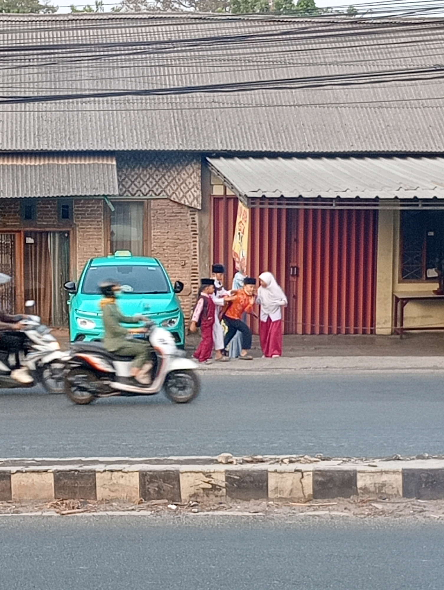 In Bandar Lampung, Indonesia, on a street, a teal-colored taxi is parked. A person on a white and orange motorcycle wearing a green uniform and a helmet with a face covering is in motion, passing the taxi. Several children are standing together on the sidewalk. Two boys are wearing white shirts, one with a red vest, and the other in an orange shirt. They are accompanied by a girl in a white hijab and a pink skirt. Behind them, a building features a textured, dark red corrugated metal gate and brick walls. The scene is bordered by a road with a striped curb in the foreground.