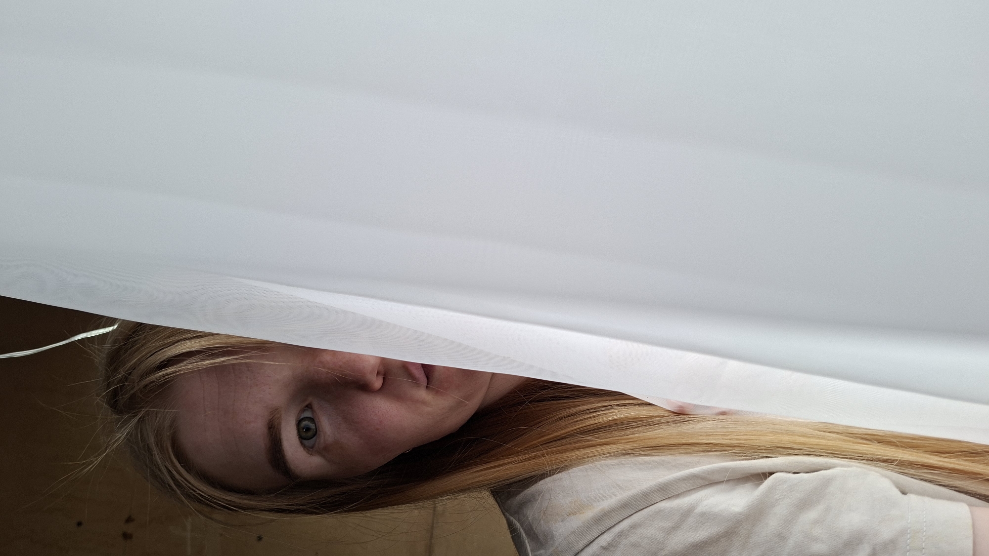 A woman with long, light brown hair and fair skin is peering out from under a white sheet or fabric. Her face is angled towards the camera, revealing her eyes and part of her nose and mouth. She is wearing a light-colored shirt. The background is a dark, textured surface. The location, Большой Букор, Russia, is not relevant to the visual content of the image.