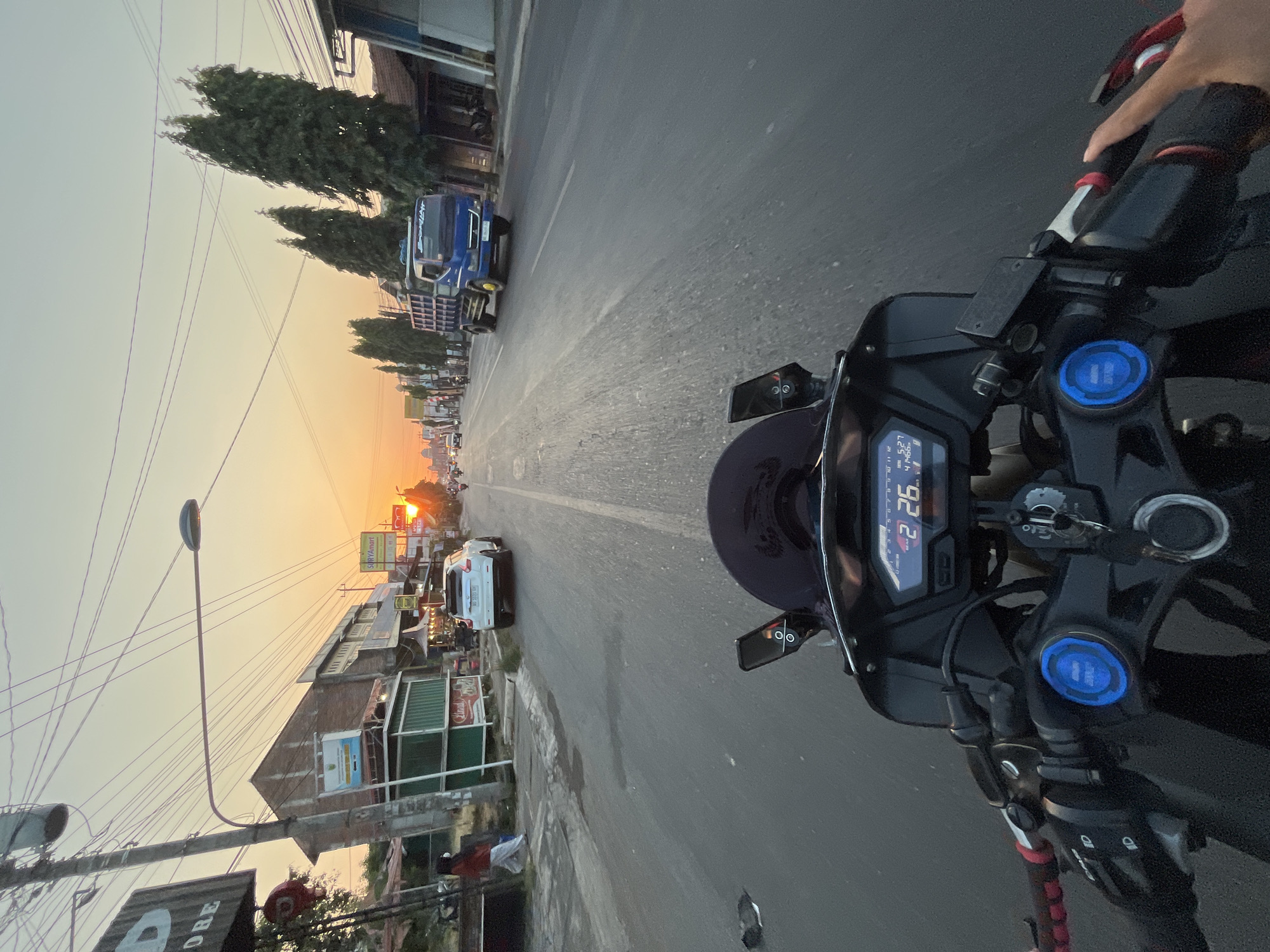 The image is taken from the perspective of someone riding a motorcycle in Kalinyamatan, Indonesia, during sunset. The motorcycle's dashboard is visible in the foreground, displaying a speed of 26 km/h. The road ahead is lined with buildings and trees, with a blue truck in the right lane. The sky is orange and yellow from the setting sun, and power lines crisscross above the street. A white car is visible on the left side of the road. A person in a red shirt and white pants is walking on the sidewalk on the left. The scene captures a typical street view in a town during the golden hour.