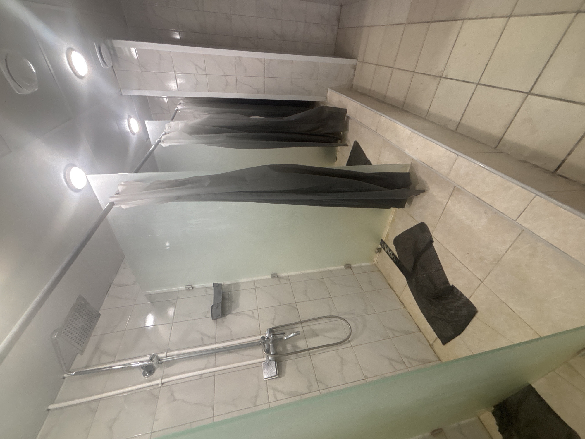 This image captures an empty, brightly lit public shower room, likely within a gym or sports facility in Perm, Russia. The scene features a row of individual shower stalls, separated by semi-transparent, light green or frosted glass partitions. Each stall is equipped with a chrome-finished wall-mounted showerhead, a mixer tap, and a flexible hose with an additional handheld shower attachment. Dark grey shower curtains are drawn across the front of some stalls, providing privacy.
The floor is tiled with light beige square tiles, exhibiting visible grout lines and some minor wear or dirt, particularly in the common walking areas outside the showers. The walls within the shower areas are covered with light-colored, possibly marble-patterned, rectangular tiles. The ceiling is white and features several circular recessed light fixtures that emit bright, cool white light, illuminating the entire space. A dark grey mat or cloth is visible on the tiled floor outside one of the shower stalls. No people are present, and no visible text can be deciphered in the image.