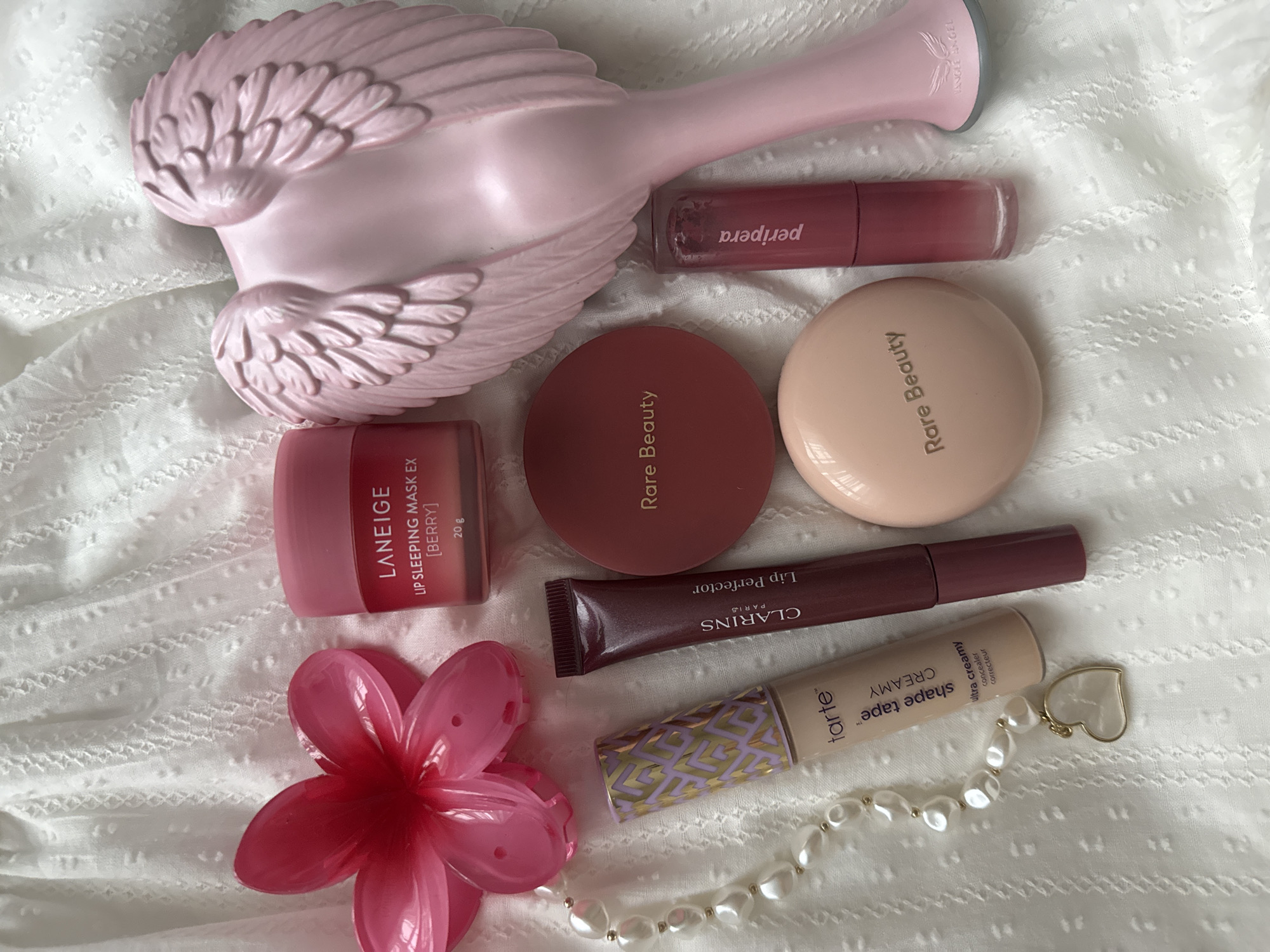 The image displays a collection of cosmetic products arranged on a white, textured fabric with a subtle pattern. The arrangement includes a pink bird-shaped object at the top left, possibly a decorative item. Below the bird, there is a jar of Laneige Lip Sleeping Mask EX, in a berry flavor. Next to the mask are two round compacts labeled "Rare Beauty." Below the Rare Beauty compacts, there is a tube of Clarins Lip Perfector, followed by a tube of Tarte Shape Tape Creamy concealer. A strand of pearl-like beads with a heart-shaped charm is placed next to the Tarte concealer. A Peripera lip product is positioned near the pink bird. A pink flower is at the bottom left corner.