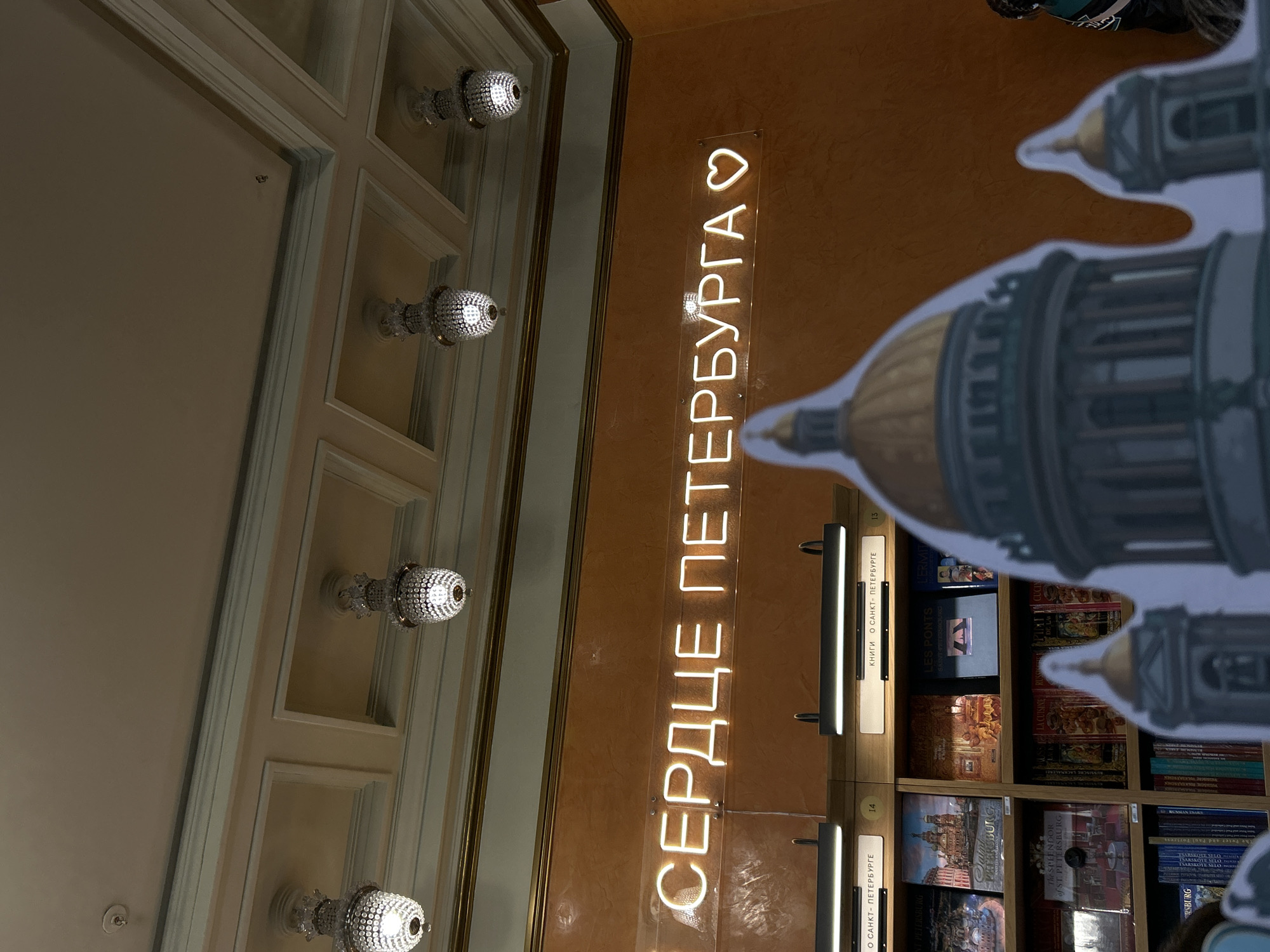 This image captures an interior scene, likely within a bookstore or souvenir shop in Saint Petersburg, Russia.
The primary subject is a vertical neon sign mounted on a textured, warm-toned wall. The sign glows white and reads "СЕРДЦЕ ПЕТЕРБУРГА♡", which translates to "HEART OF PETERSBURG♡". To the left, a portion of an elaborately decorated, light-colored ceiling is visible, featuring recessed panels and multiple ornate, crystal-like light fixtures emitting a bright glow.
To the right of the neon sign, bookshelves are packed with books. Several titles are visible, suggesting a theme related to Saint Petersburg and Russia. Visible English titles include "The SPLENDOR of ST. PETERSBURG", "LES PONTS SAINT PETERSBOURG" (The Bridges of Saint Petersburg), "LERMIT" (likely The Hermitage), and a partial "TSARSKOYE SELO". Russian text includes "КНИГИ О САНКТ-ПЕТЕРБУРГЕ" (Books about Saint Petersburg) on shelf dividers numbered "13" and "14". A large, light-blue cardboard cutout, shaped like a prominent architectural structure resembling St. Isaac's Cathedral, partially obstructs the view of the bookshelves on the right.
The overall setting emphasizes a dedication to the city of Saint Petersburg, reinforced by the visible text and architectural decorations. The lighting appears to be artificial, typical of an indoor commercial space. No people are clearly visible in the frame, indicating a static display of the shop's interior.