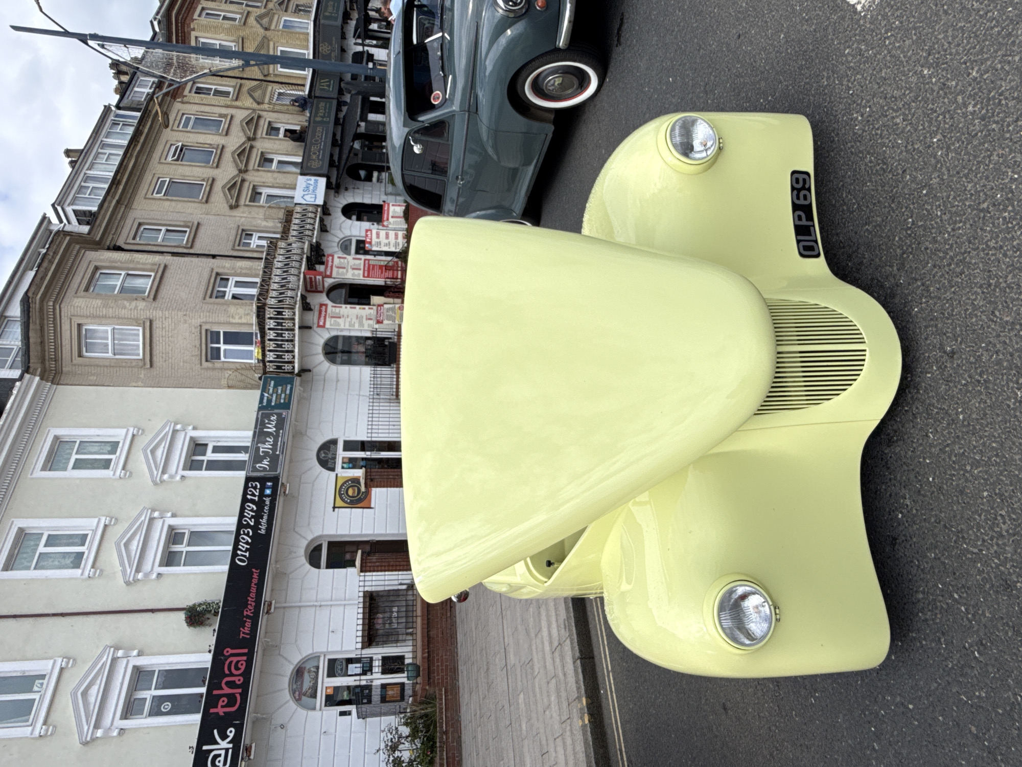 Here is a detailed description of the image:
This outdoor shot, taken at street level, features the front of a light yellow vintage car with the license plate "OLP 69". The car is parked on an asphalt road and is angled towards the right side of the frame. A second, dark-colored classic car is visible behind it.
The backdrop of the image reveals an urban landscape. A multi-story building with off-white façade is situated behind the cars. "ek, thai" is displayed in a prominent sign "Thai Restaurant", accompanied by the phone number "01493 249 123" and the words "In The Mix". "Sky's House" and "HOTEL Ocean" are also visible on signs. Based on the style of architecture and signage, this photograph likely was taken in Great Yarmouth, United Kingdom. The building and other businesses suggest a commercial area of a city.