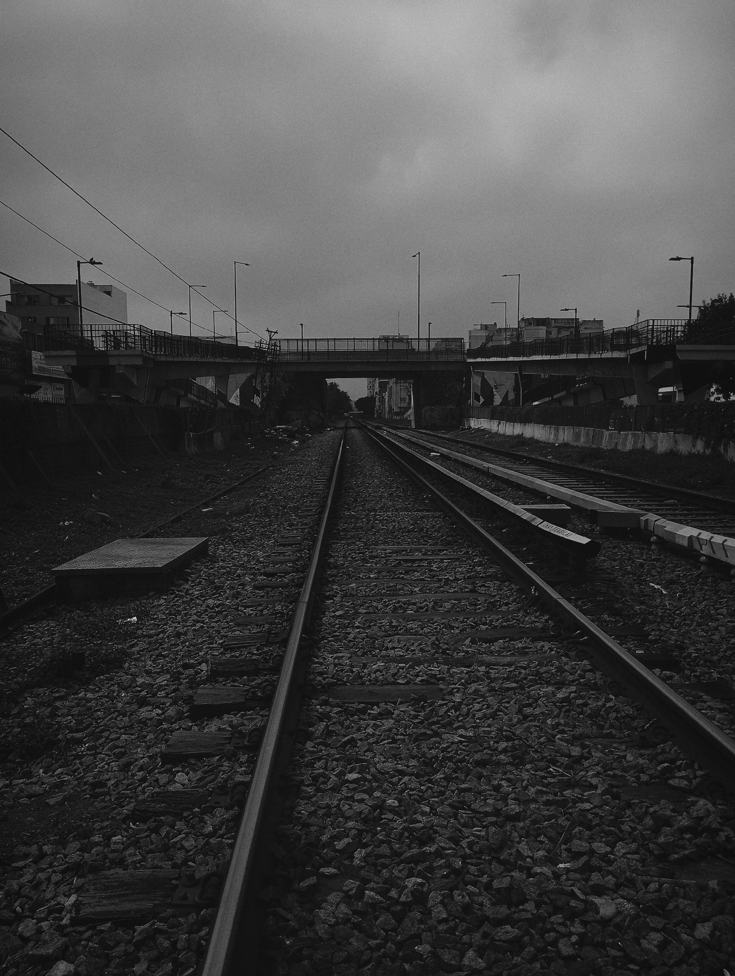 The image shows a railway track running towards a bridge in Buenos Aires, Argentina. The tracks are surrounded by ballast. Above the tracks is a road overpass. Streetlights and buildings are visible along the overpass. The sky is overcast. The image is in black and white.