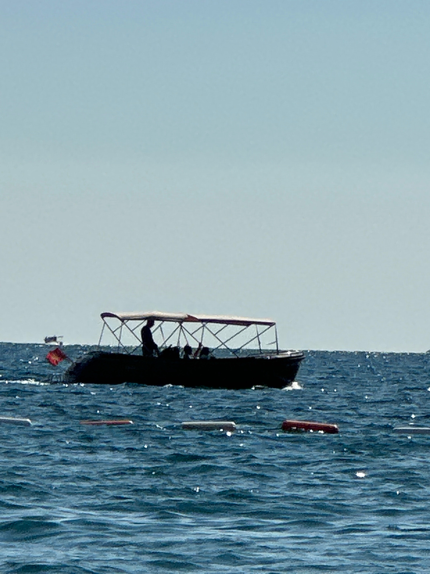 An eye-level, medium shot captures a wide view of the Adriatic Sea off the coast of Boreti, Montenegro, under a clear, pale blue sky. The water is a deep blue with whitecaps and shimmering reflections of sunlight. In the mid-ground, a dark-hulled boat with a beige canopy travels from left to right. The boat is carrying several silhouetted figures, including one standing near the front who appears to be operating the vessel. To the left of the boat, a small red flag flutters on a pole. In the foreground, several cylindrical buoys are partially submerged in the water, marking a boundary or path. The scene is peaceful and evokes a sense of calm summer weather.