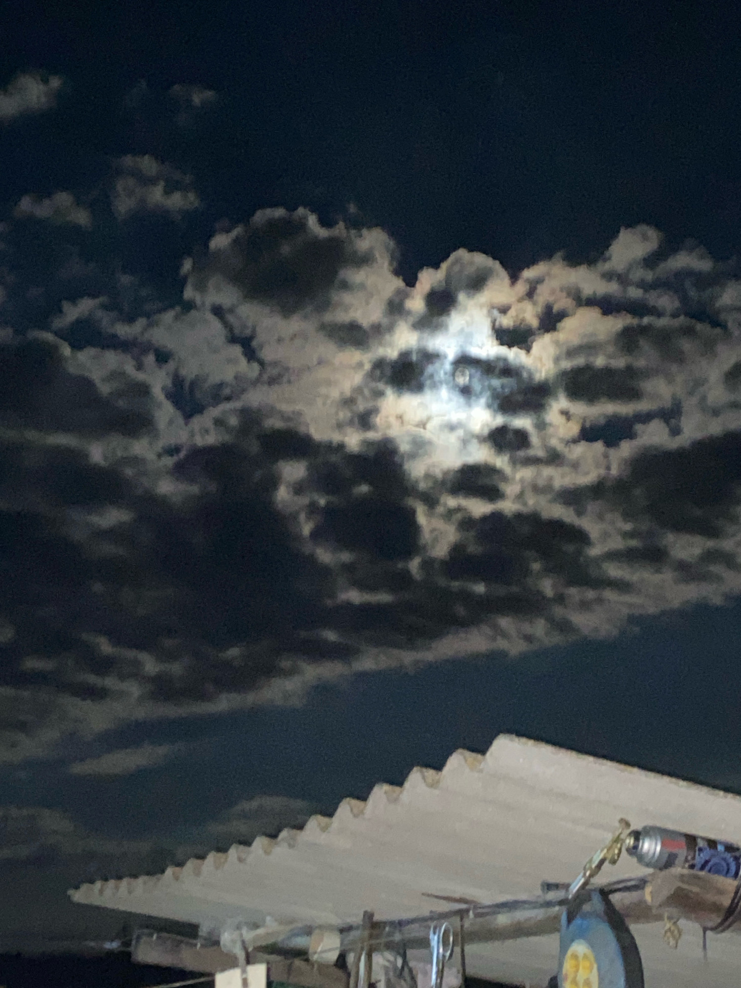 A low-angle, nighttime photograph captures the moon peeking through a cloudy sky, illuminating a corrugated roof in the foreground. The moon appears as a bright, diffused orb amidst dark, textured clouds. The roof, likely made of asbestos cement sheeting, dominates the lower portion of the frame, with its distinctive wavy pattern creating a sharp diagonal line from the lower left to the upper right. A few objects are visible on or near the roof, including a silver-colored aerosol can with blue markings and a pair of scissors hanging from a hook. The background consists of a dark, deep blue night sky filled with scattered clouds. The overall scene suggests an outdoor environment, possibly a rooftop or an elevated vantage point, in an unlit urban or rural setting, likely the city of Besplemyanovskii, Russia, given the context.