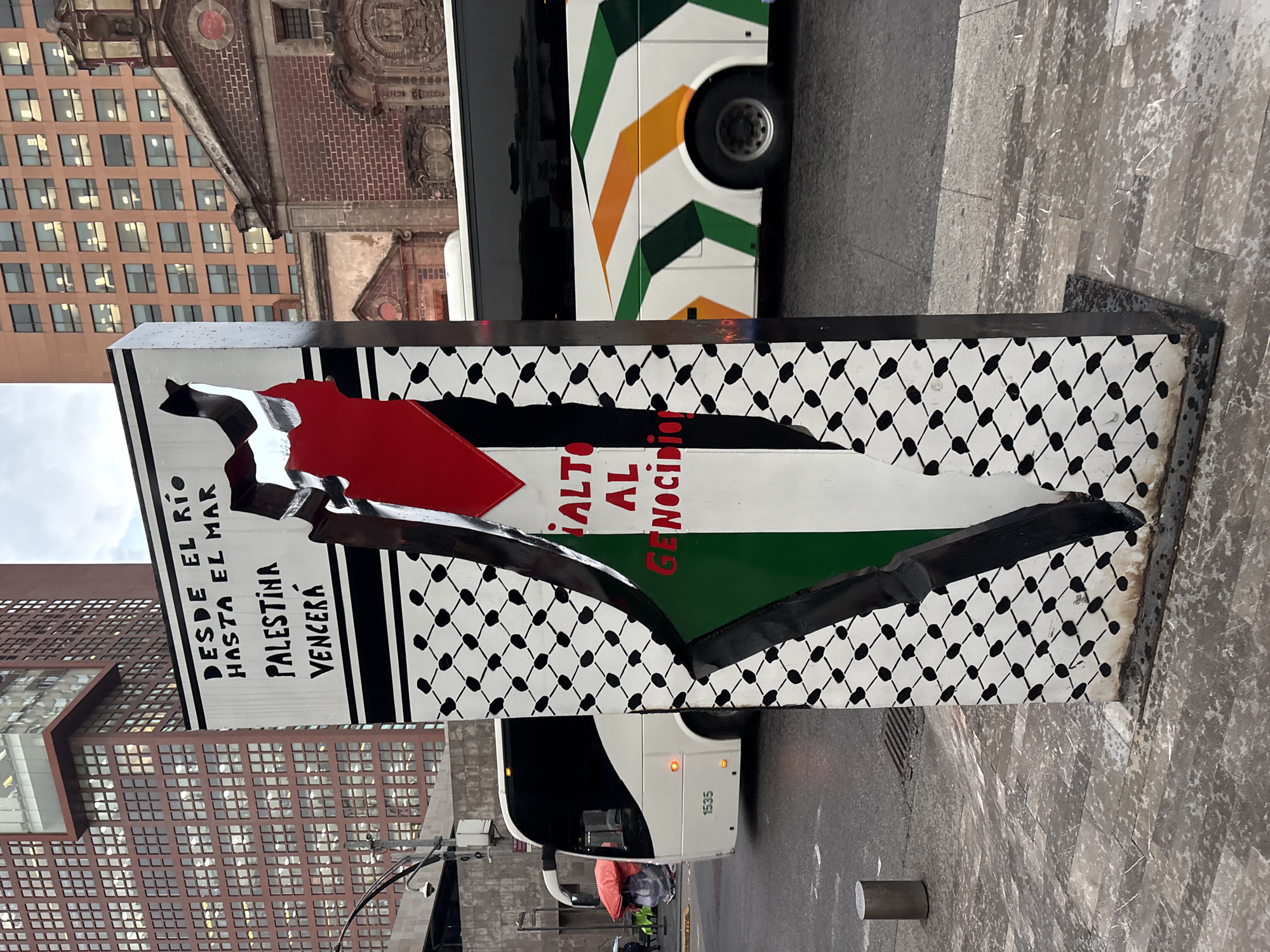 This is a street-level, eye-level, full-shot photograph taken in Mexico City, Mexico. The primary subject is a tall, rectangular monument with a map of Palestine overlaid with the Palestinian flag colors (red, white, black, and green). The monument is adorned with text in Spanish, reading "DESDE EL RÍO HASTA EL MAR, PALESTINA VENCERÁ" which translates to "From the River to the Sea, Palestine will prevail" and "¡ALTO AL GENOCIDIO!" which translates to "Stop the Genocide!". The monument is positioned on a sidewalk, with a busy street visible in the background. A white tour bus with green and orange stripes is driving past the monument. In the background, there are tall buildings, one with a red brick facade and many windows, and another modern building with a dark facade. The sky is overcast. The ground is paved with stone tiles and asphalt.