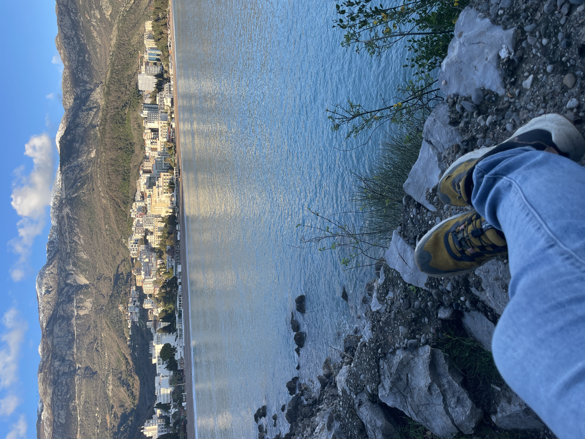 The image displays a coastal landscape in Budva, Montenegro, captured from a low, first-person perspective. In the immediate foreground, the lower legs and feet of an individual are visible, wearing light blue denim jeans and dark yellow/olive athletic shoes with prominent black accents and white soles. The person appears to be seated on or near a rocky embankment composed of large, grey boulders interspersed with smaller gravel and sparse green vegetation, including small shrubs with yellow flowers.
The midground features the calm, blue-green waters of a bay, showing small ripples and reflections of sunlight. Along the immediate shoreline in the foreground, dark rocks are partially submerged, with gentle waves lapping against them.
Across the bay, the city of Budva is prominently displayed. A long, light-colored sandy beach lines the water, backed by numerous multi-story buildings, ranging from hotels and resorts to residential structures, primarily in white and light earth tones. Scattered green trees and vegetation are visible among the buildings.
The background is dominated by large, rugged mountains rising directly behind the cityscape. These mountains exhibit exposed rock faces and sparse brown vegetation, with patches of snow visible on their higher peaks, indicating a cooler climate or season. The sky above is a clear blue with scattered white cumulus clouds. The lighting suggests either early morning or late afternoon, with sunlight reflecting off the water and illuminating the city and mountain slopes.