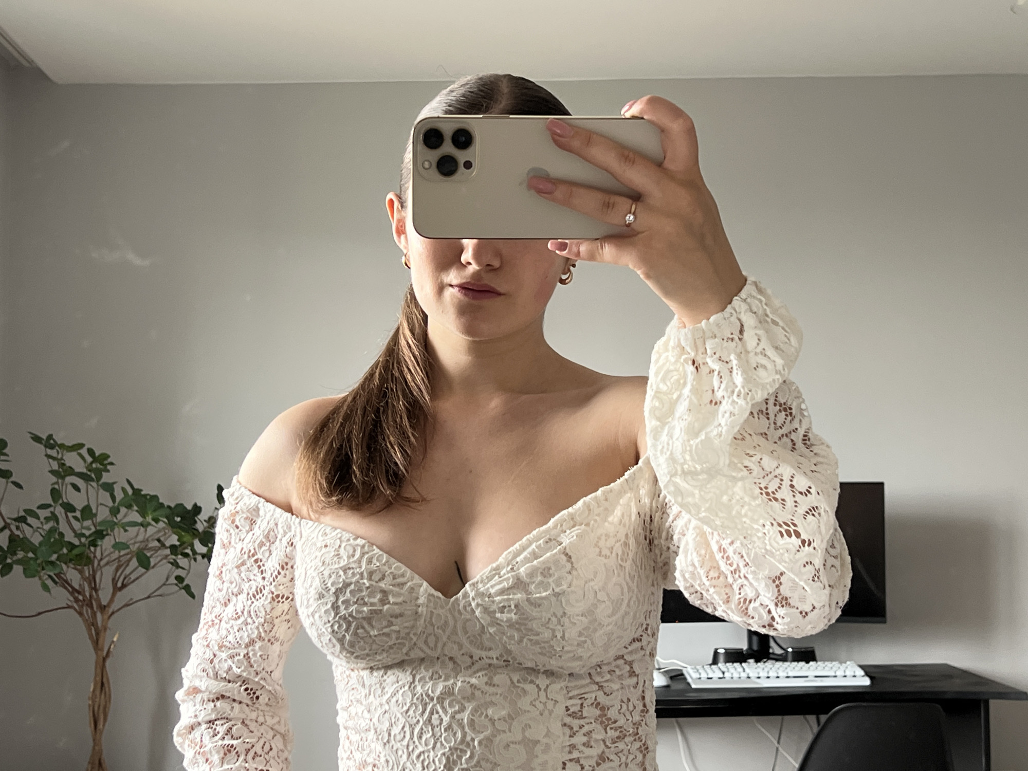 The image displays a young woman taking a mirror selfie indoors, likely in a room located in Ternopil, Ukraine. She holds a light-colored smartphone, possibly an iPhone due to its triple camera setup, horizontally in front of her face, effectively obscuring her eyes and forehead. Her dark brown hair is pulled back, with a low ponytail visible over her left shoulder. She wears a cream or white off-the-shoulder top or dress made of intricate lace with a floral pattern, featuring long, voluminous sleeves gathered at the wrists. A delicate ring is visible on her right hand's ring finger, and she has small hoop earrings. A subtle mark or tattoo is visible in her cleavage.
The setting appears to be a modern indoor space with light grey walls and a white ceiling. To the left, a small potted plant with green leaves and a thin trunk adds a touch of greenery. On the right, a dark desk is equipped with a black computer monitor, a white keyboard, and a dark office chair, suggesting a home office or personal workspace. The room is well-lit by natural light, indicating it is daytime. The woman's visible mouth suggests a neutral or slightly relaxed expression as she captures her reflection.