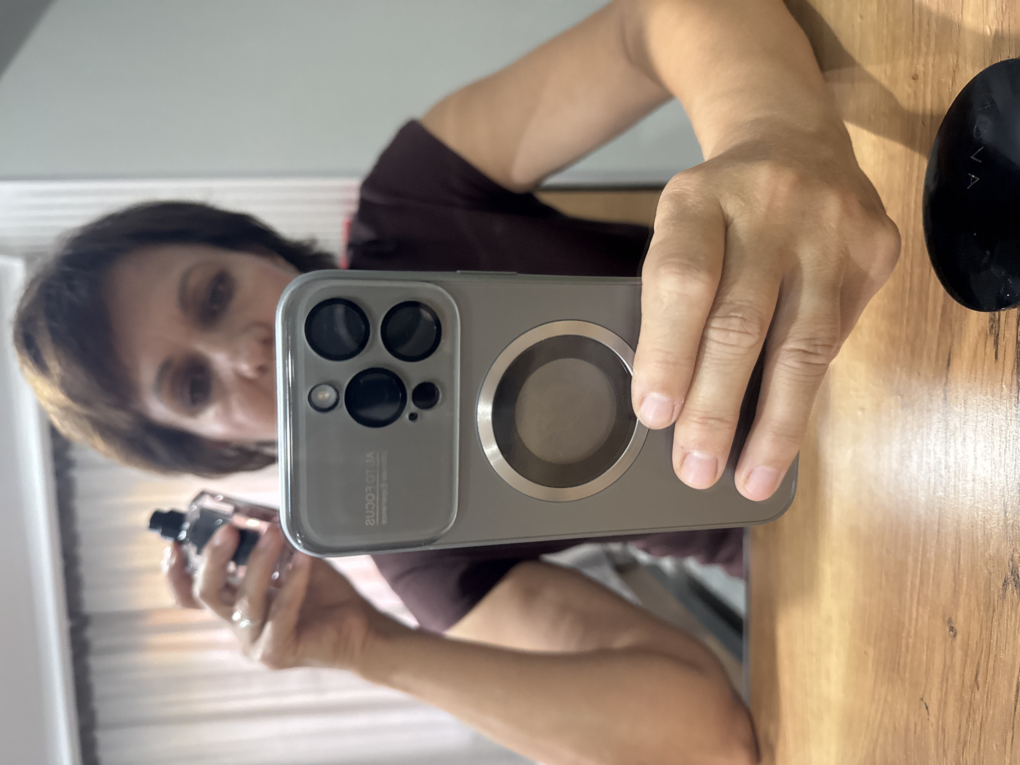 A woman is taking a selfie in an indoor setting. She is holding a smartphone in her right hand, angled to capture her reflection. In her left hand, she holds a dark-colored bottle, possibly perfume or a cosmetic product. The woman has short, dark brown hair and is wearing a maroon t-shirt. She is seated at a wooden table. In the background, sheer curtains are visible. The image is cropped to focus on the woman's upper body and hands. There is a black, oval-shaped object on the table in the foreground, with some faint lettering on it. The overall scene appears to be a personal moment, possibly at home. The location of Солонешное, Russia is not determinable from the visual information provided in the image.