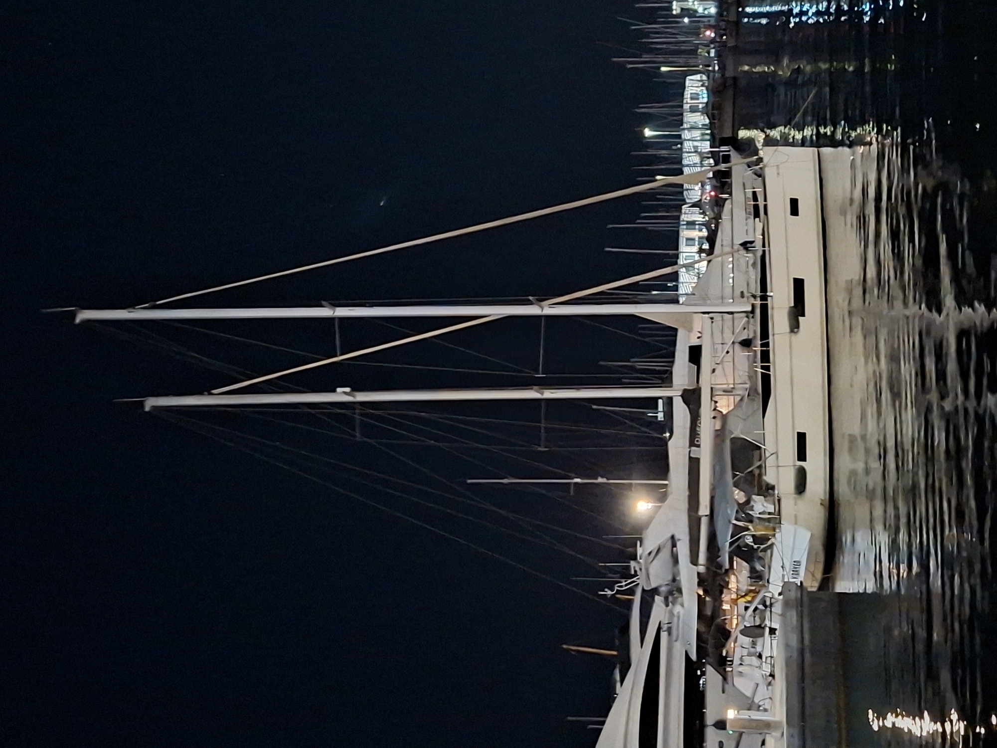 This is a nighttime, medium-range, eye-level, full-frame shot of yachts docked in a marina. The dominant subjects are two large sailboats, one in the foreground and one slightly behind it and to the left. The foreground sailboat is white with a gray canopy over the cockpit and has "KRAKEN" written in black lettering on its stern. The sailboat behind it is larger and whiter, with a sleeker design. Numerous other masts and rigging of other sailboats are visible in the background, creating a dense, vertical forest of poles. The water in the foreground reflects the lights from the marina and the boats, creating streaks of white and blue. The sky is dark and clear, with no visible celestial bodies. The overall impression is of a quiet, well-lit harbor at night. The yachts appear to be in the marina of Alicante, Spain, given the style and type of vessels, commonly found in Mediterranean ports.