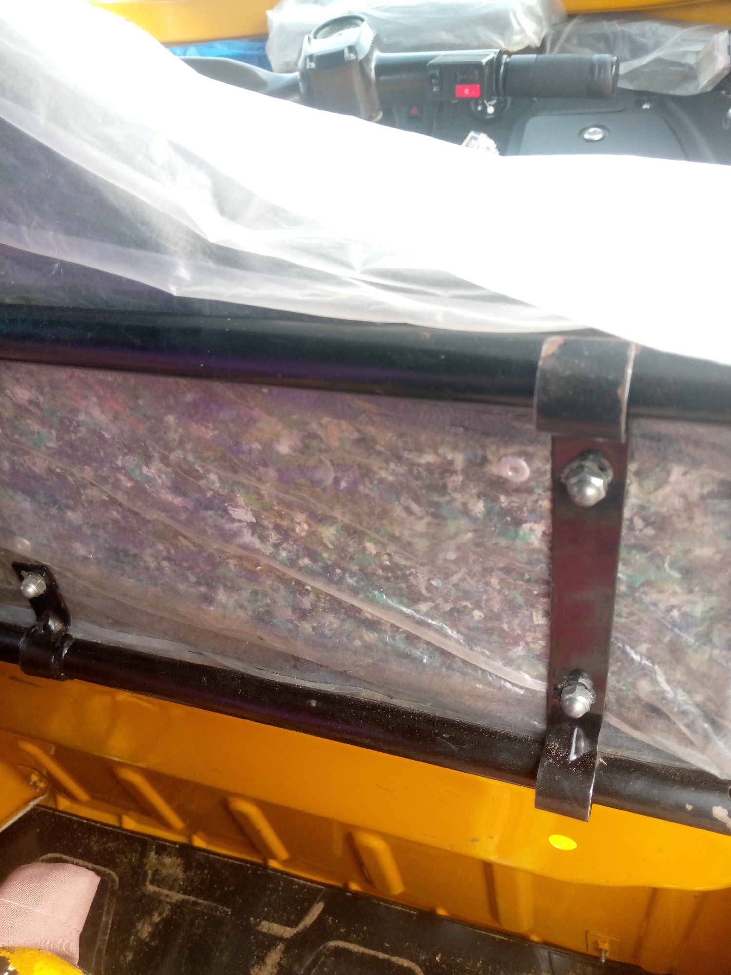 The image shows a close-up view of the interior of a yellow vehicle, likely a three-wheeled auto-rickshaw, in Jalingo, Nigeria. The focus is on the passenger area, which is covered with a transparent plastic sheet. Several metal brackets and bolts are visible, securing the plastic cover to a black metal frame. The vehicle's dashboard, including a handlebar and controls, is partially visible at the top of the image, covered with a white plastic sheet. The overall scene suggests the vehicle is either new and being protected or has been modified with this covering. The environment appears to be a workshop or storage area, given the protective sheeting and the utilitarian nature of the vehicle.