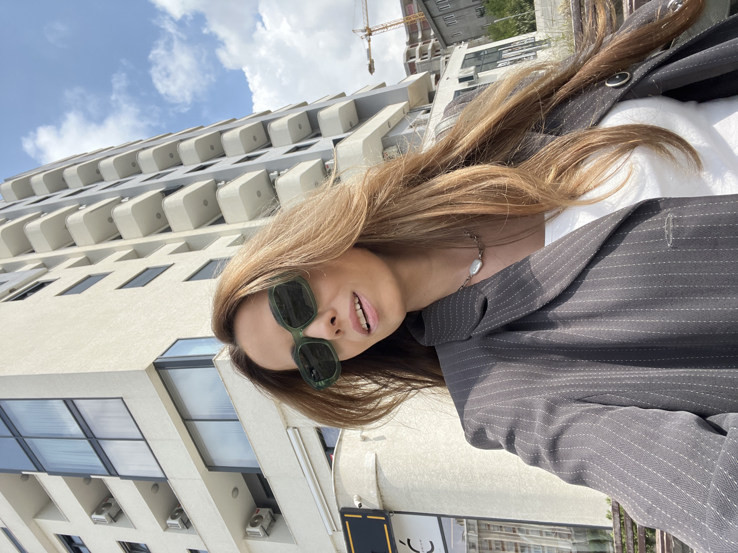 A bright, daytime outdoor selfie captures a woman smiling and looking towards the camera, set against a modern urban backdrop. The woman has long, light brown hair and wears dark green, rectangular-framed sunglasses. She is dressed in a dark grey pinstriped blazer over a white top, accessorized with a silver chain necklace featuring an irregular white pearl-like pendant.
The background is dominated by a tall, light-colored modern building characterized by multiple identical, rounded greyish-white balconies or architectural protrusions stacked vertically. Below these, dark-paned windows are visible, and several white air conditioning units are mounted on the lower levels of the building. The sky is bright blue with scattered white clouds, indicating clear and sunny weather. A construction crane is partially visible in the upper right background, suggesting development in the area.
In the bottom left corner, a dark sign is partially visible, displaying the text "DIAM" and "PREMIUM" along with a stylized crescent-shaped logo. The image conveys a cheerful and contemporary urban scene.