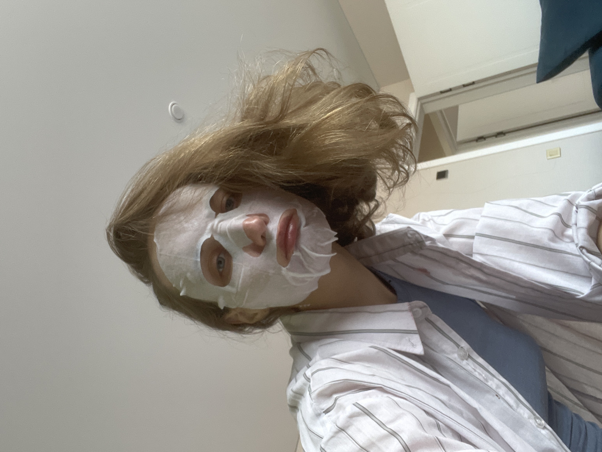 The image depicts a person lying down, looking upwards at the camera. They are wearing a white sheet mask on their face. The person has wavy blonde hair. They are also wearing a white and gray striped button-down shirt over a blue top. The background includes a white ceiling and a white cabinet or window frame. It is not possible to confirm the location in the city of Sepych, Russia based solely on the visual information provided.