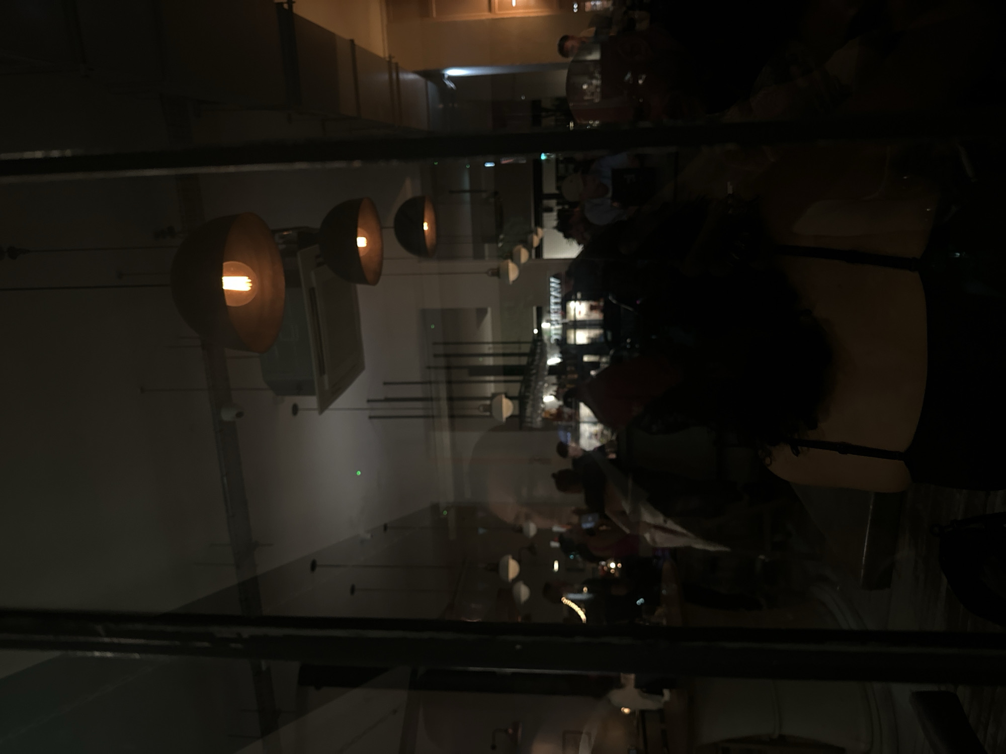 A dark, reflective image taken through a window or glass panel depicts the interior of a restaurant in London, United Kingdom. In the foreground, the frame of the window or glass panel is visible. The interior of the restaurant is dimly lit with industrial-style pendant lights hanging from the ceiling, casting a warm glow. Several tables are occupied by patrons, suggesting a lively atmosphere. The reflection adds a layer of complexity, creating a sense of depth. Details such as the bar area and silhouettes of people are discernible in the background. The overall ambiance suggests a trendy or modern dining establishment.