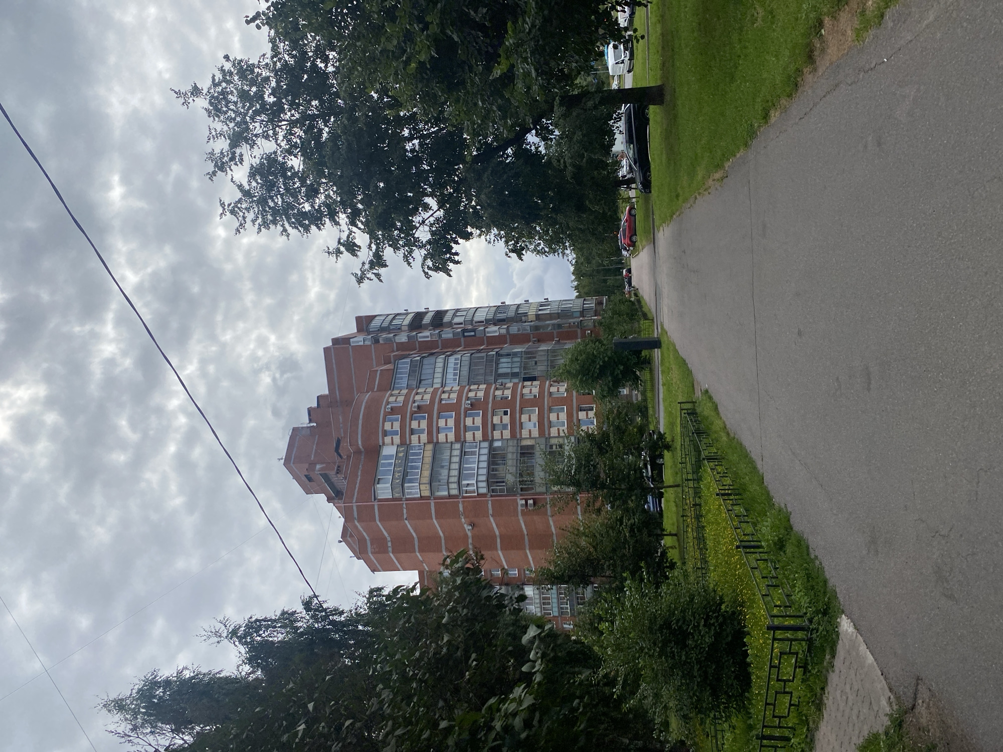 An overcast day in Saint Petersburg, Russia, presents a view of a tall, multi-story, brick residential building with numerous balconies and windows. The building's facade is a warm, reddish-brown brick, accented with lighter horizontal bands. Adjacent to the building, a paved walkway curves gently to the right, bordered on the left by a low, black metal fence enclosing a patch of grass dotted with yellow flowers. Several trees with dense green foliage are scattered throughout the scene, with some framing the building and others lining the walkway. On the right side of the walkway, a grassy area with a mature tree is visible. In the background, cars are parked along the side of a road. The sky is filled with thick, gray clouds, suggesting a potential for rain.